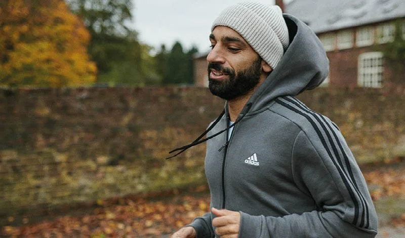 A man jogging outdoors in a gray Adidas hoodie with black stripes, wearing a light gray beanie, on an autumn day with fallen leaves and a brick wall in the background.