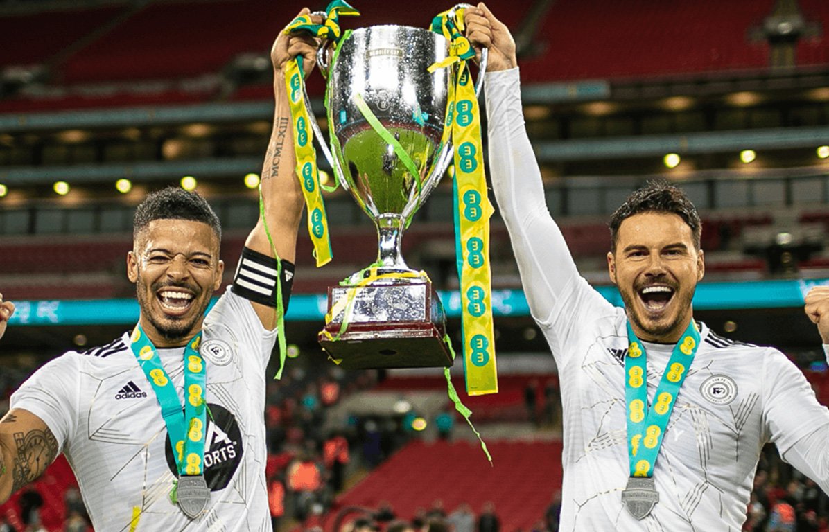 Two soccer players wearing white jerseys and medals around their necks celebrate on the field, holding a large silver trophy together with yellow and green ribbons.