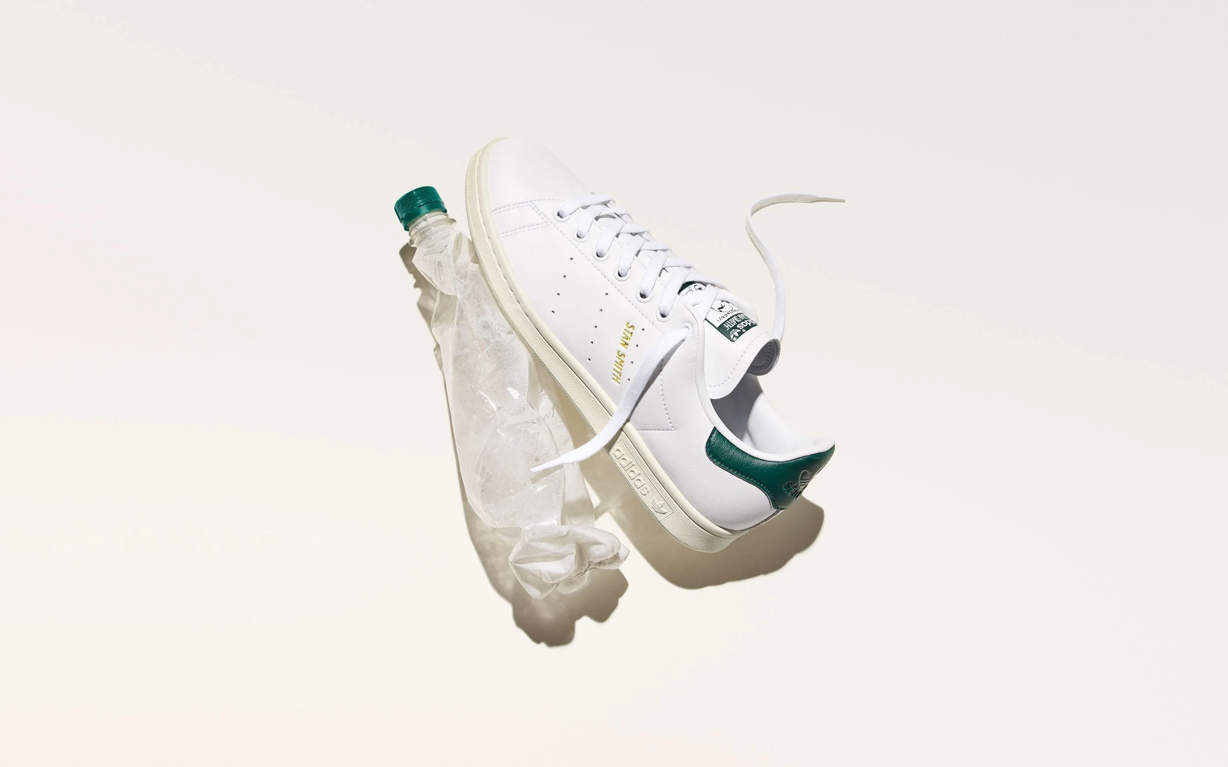 A white sneaker with green accents, a green heel tab, and the words 'STAN SMITH' printed on the side, is hanging above a crumpled plastic water bottle with a green cap on a beige background.
