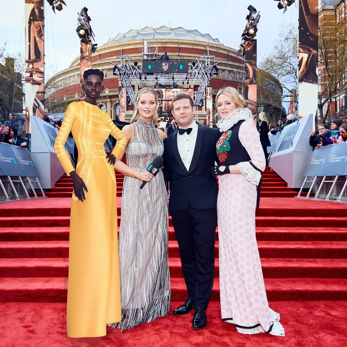 Four people standing on a red carpet outside a building with historical architecture, surrounded by crowd and photographers, during an event. The group includes a woman in a yellow dress, a woman in a metallic striped gown holding a microphone, a man in a tuxedo, and a woman in a pink and black embroidered dress with a lace collar.