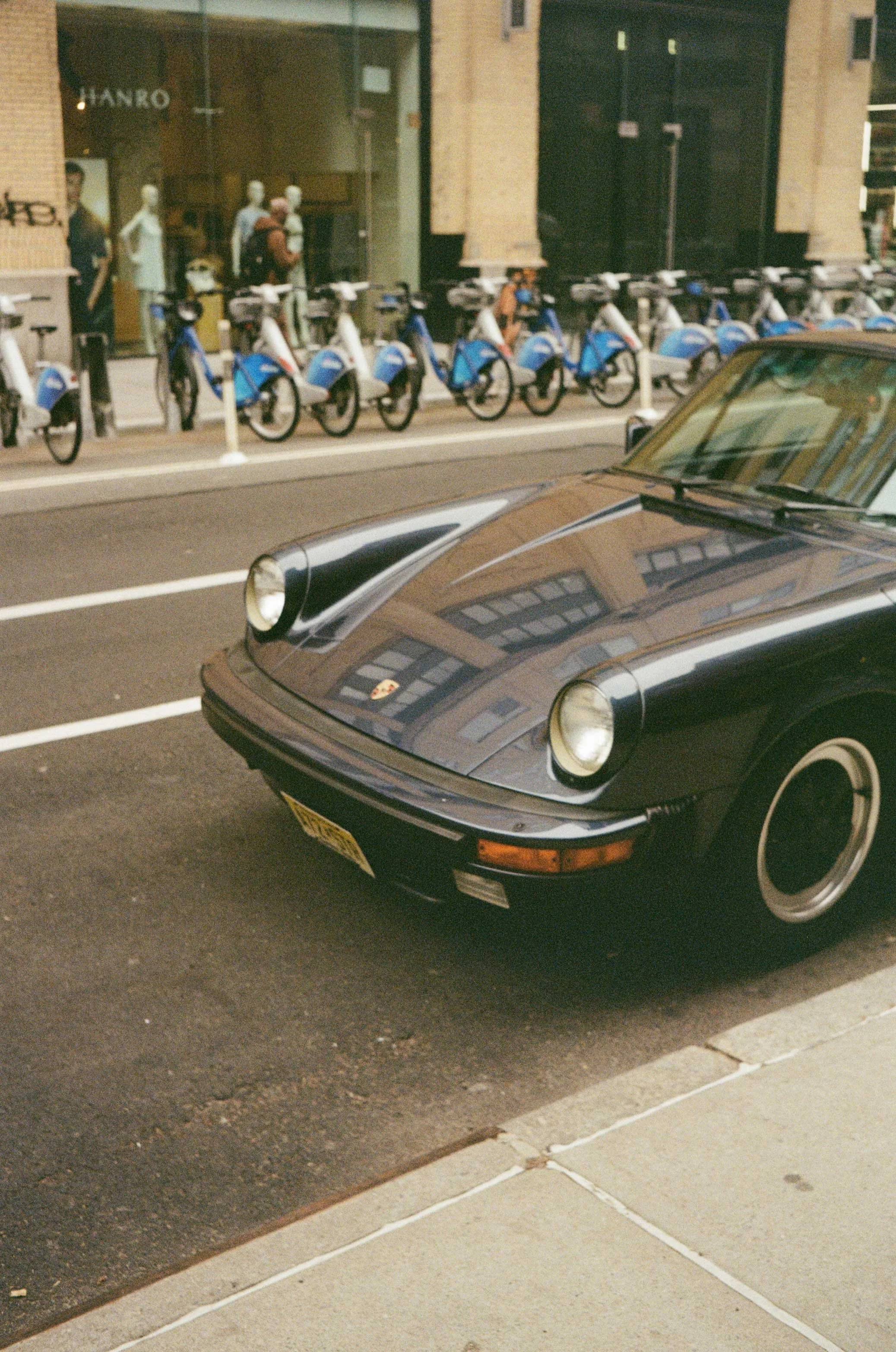 Close-up of a black Porsche car parked on the street with a row of blue and white bike share bikes parked at a bike rack on the sidewalk in the background, with a storefront and pedestrians visible.