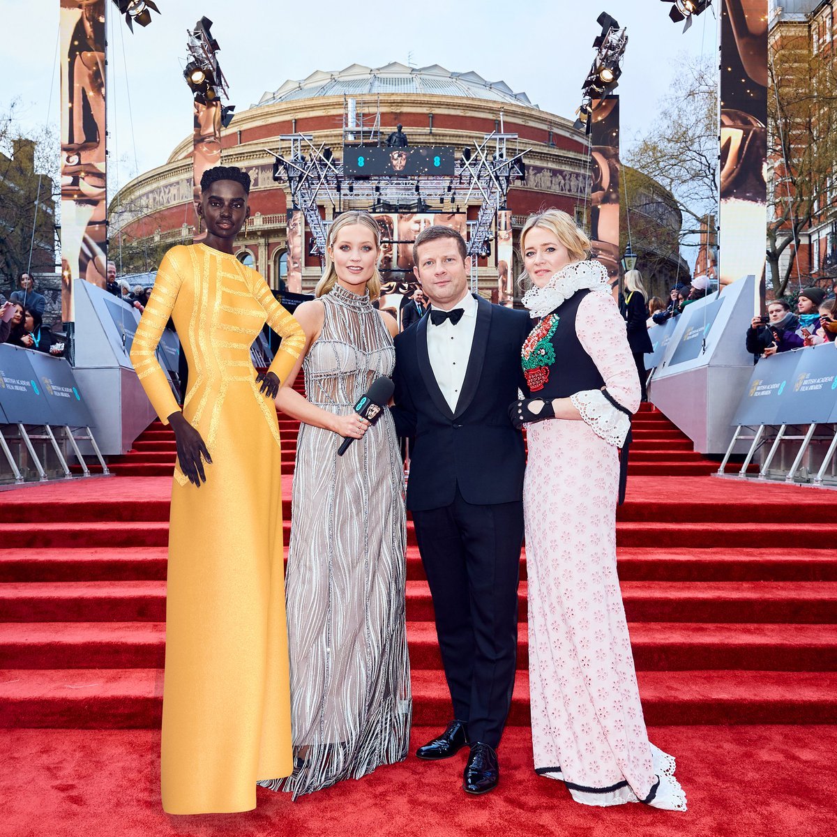 Four models and a man in a tuxedo standing on a red carpet outside a building with a grand domed roof, surrounded by media and spectators, at a formal event or award ceremony.