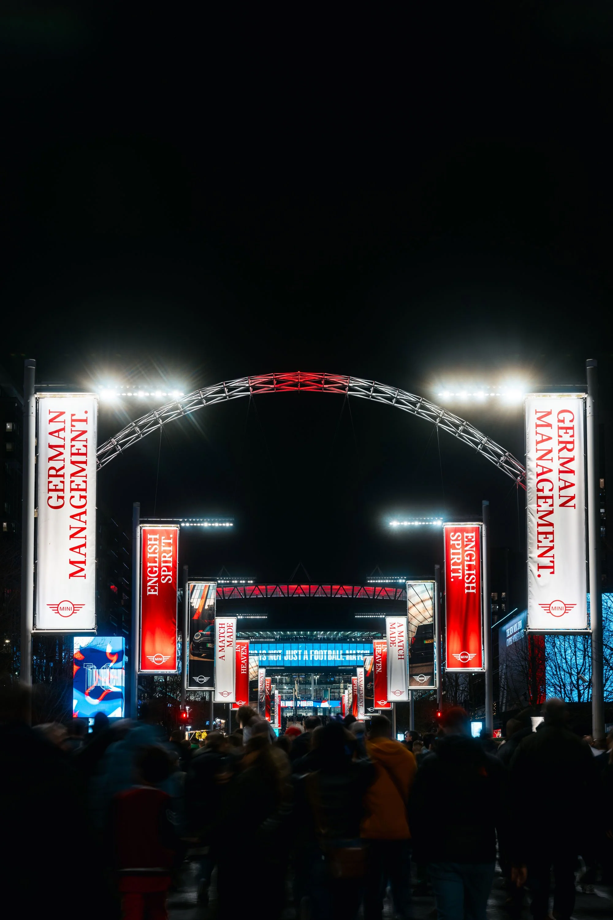 Nighttime scene at a crowded outdoor event with illuminated banners and a large screen, people walking underneath a lit archway.