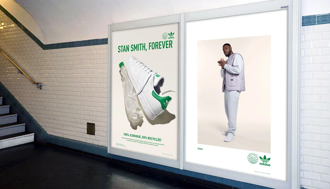 Advertisement in a subway station featuring a white Adidas sneaker with a green heel tab, a plastic bag, and a green bottle, with the text 'Stan Smith, Forever' and statements about recycling and being iconic. Next to it, a man in a grey jacket, white pants, and white sneakers stands with his hands clasped.