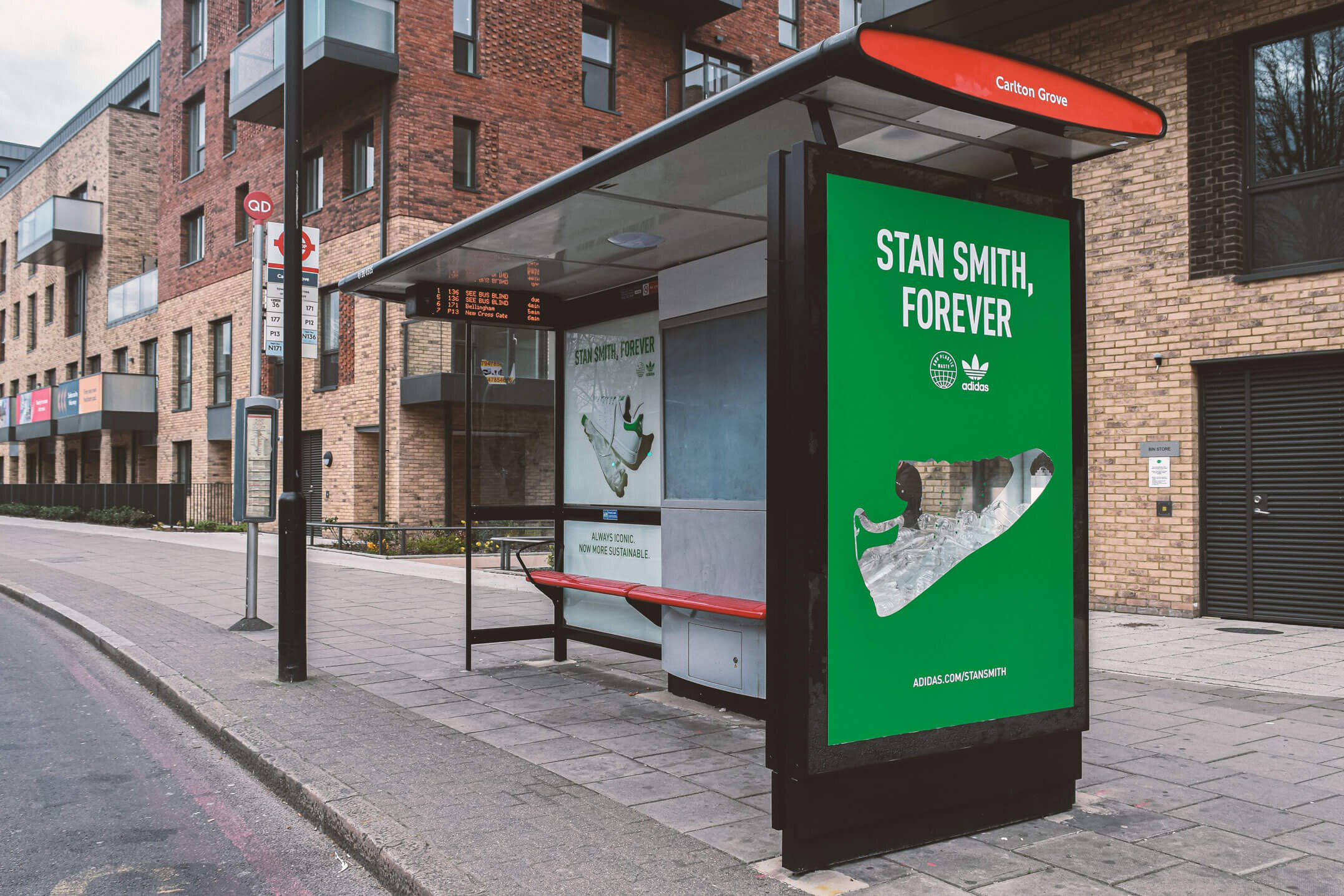 A bus stop shelter on a city sidewalk with a green advertisement for Adidas Saint Smith, Forever, showing an ice cream cone, and a digital bus schedule display. In the background, there are brick residential buildings and a street with no vehicles or pedestrians.