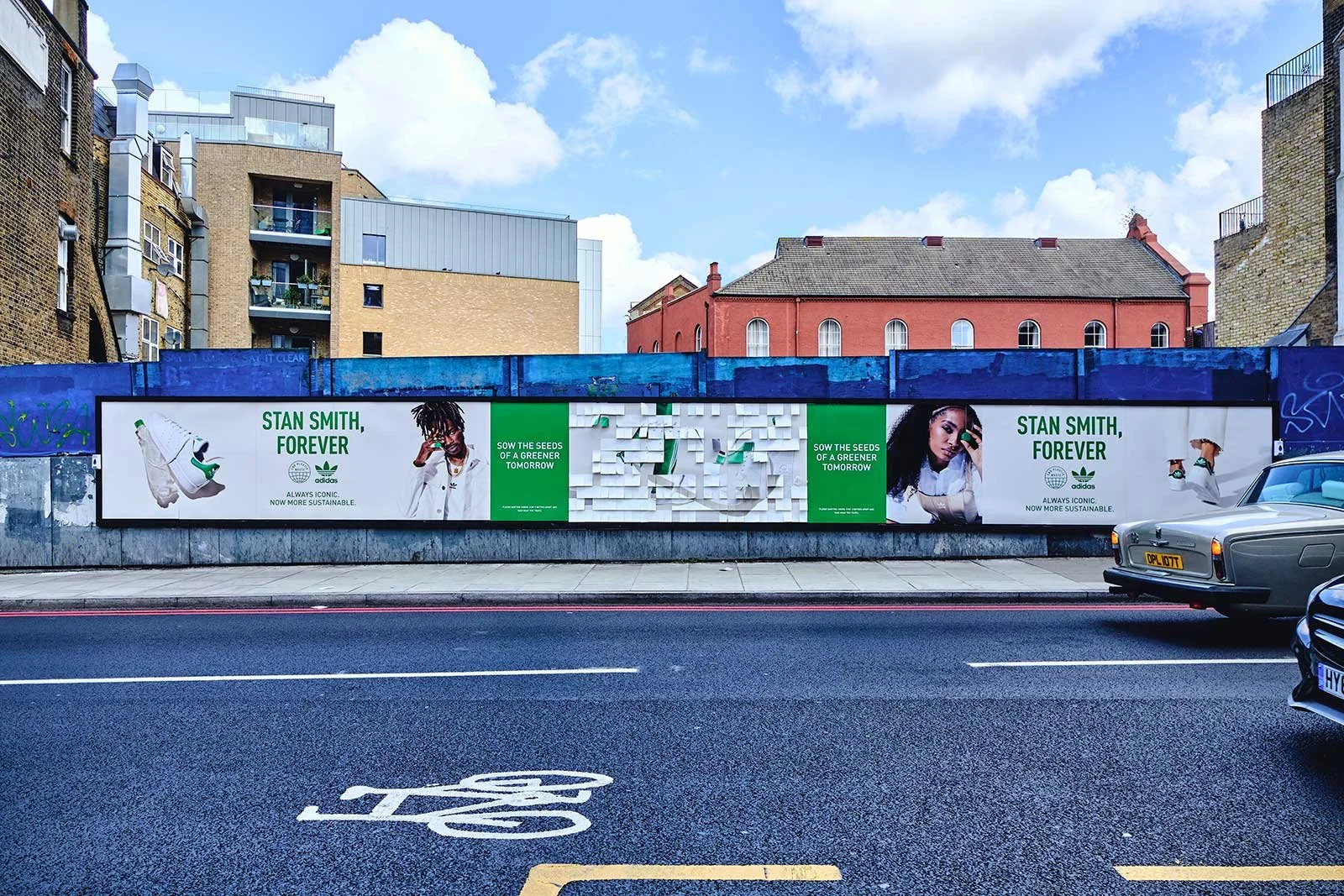 Billboard advertisement for Adidas featuring images of a man and a woman, with green and white text, set against a blue wall in an urban area, with cars on the street in the foreground and buildings in the background.