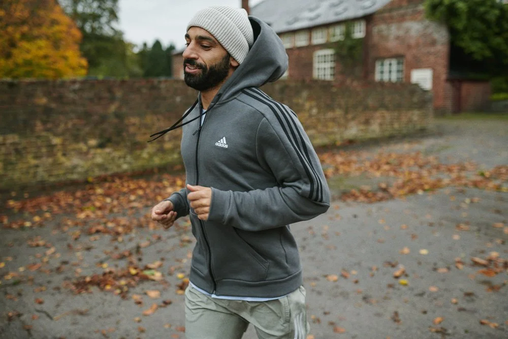 A man jogging outdoors on a fall day, wearing a gray hoodie, gray beanie, and beige pants, with autumn leaves on the ground and a brick building in the background.