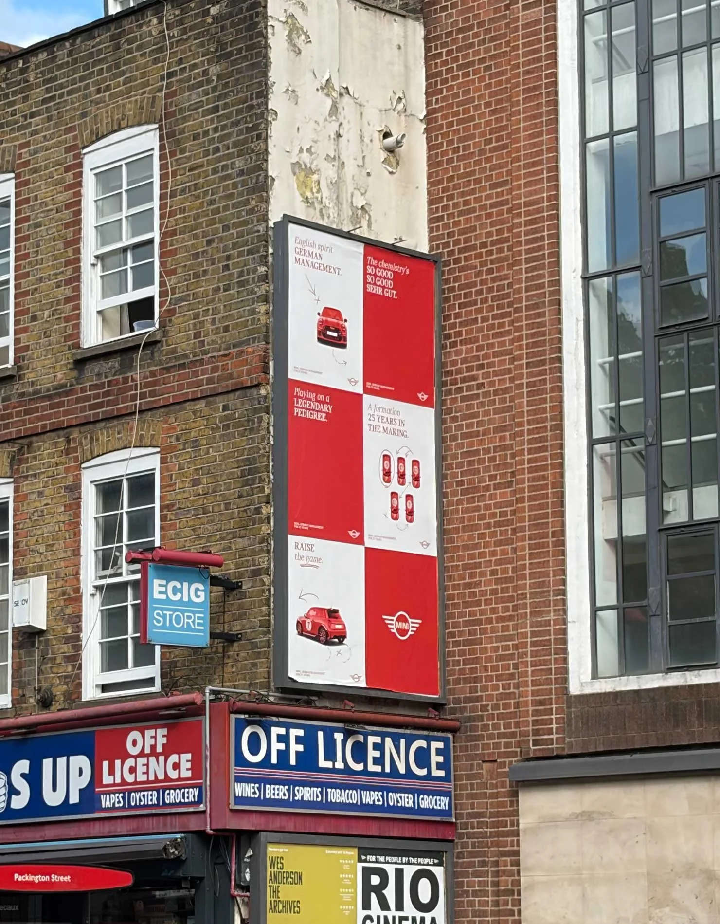 A city street scene showing a multi-story brick building with a large vertical advertisement for Mini cars. The ad features images of a red Mini car and various promotional text. Below the ad, there are signs for an off license store offering vapes, oyster, grocery, wine, beers, spirits, tobacco, and more. There is also a blue sign for an ECIG store and a small yellow sign for Rio Cinema.