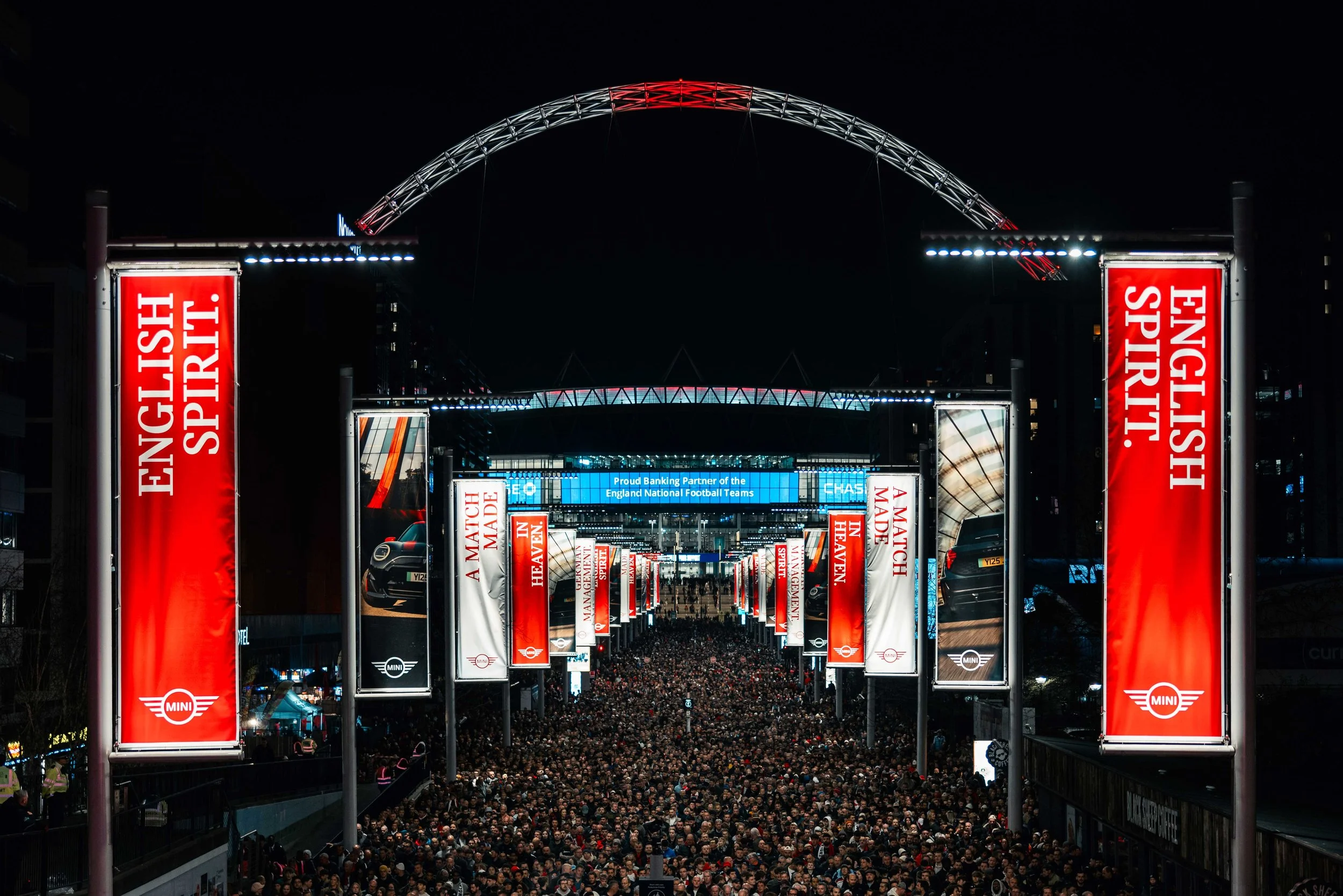 Night scene of a large crowd gathered at an outdoor event with tall red banners and illuminated signs, including the Mini logo, and a large modern arch structure overhead.
