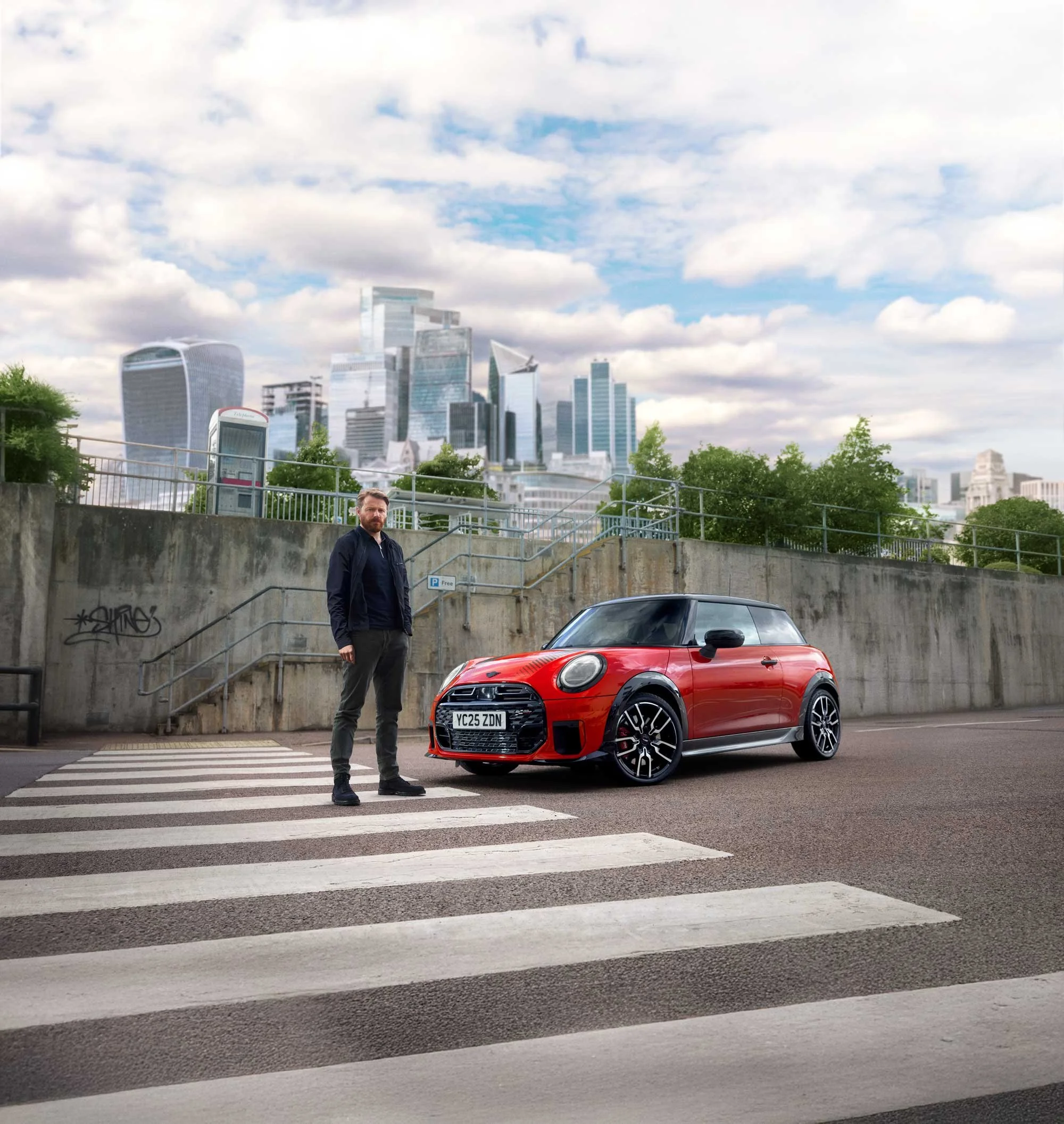 A man with a beard in dark clothing stands next to a red Mini Cooper car at a pedestrian crosswalk with a city skyline of modern skyscrapers in the background.