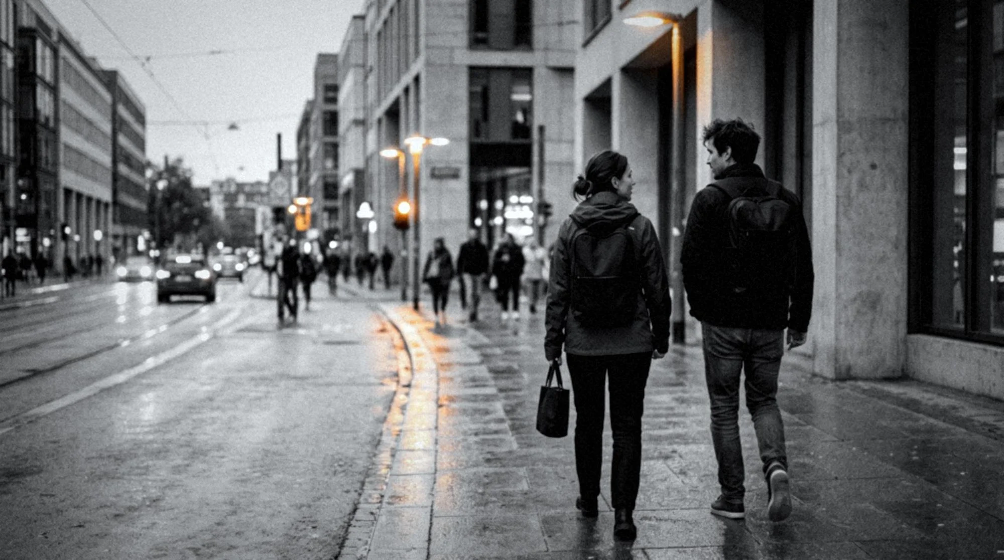 Two people with backpacks walking along a rainy urban sidewalk beside a busy city street at dusk