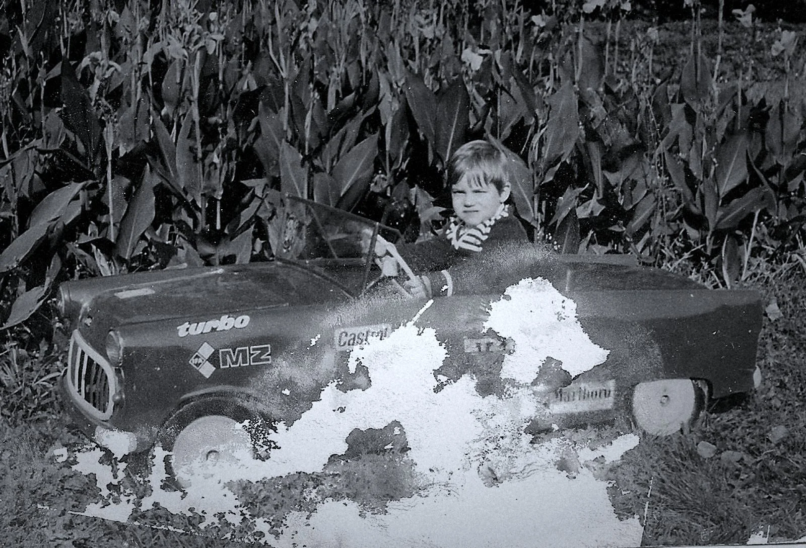 Black-and-white vintage photo of a child driving a miniature EMZ Turbo tractor through a garden.