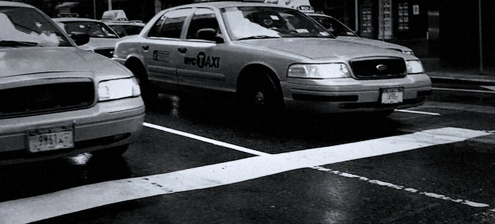 Black-and-white photo of parked city taxis on a wet street, including a Ford sedan marked “TAXI.”