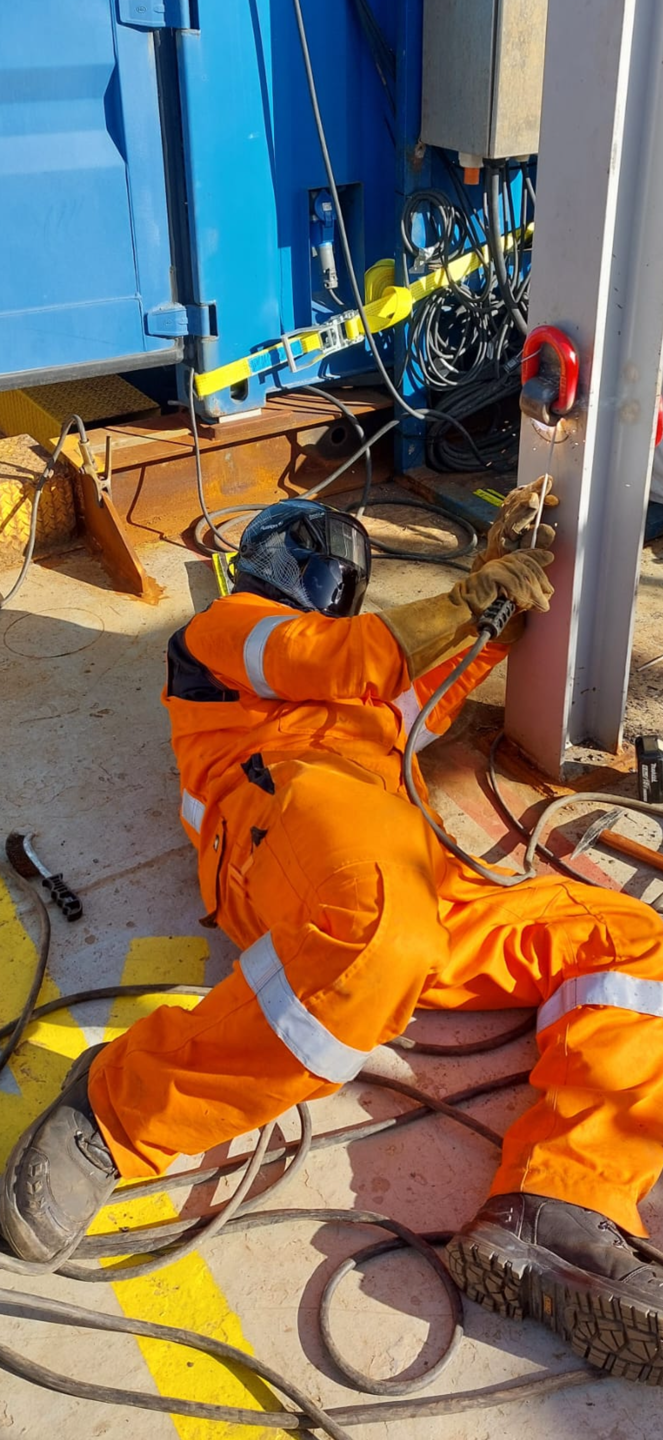 Worker lying on ground in orange safety gear, welding or working on a metal post with electrical tools and hoses around, outdoor construction site.