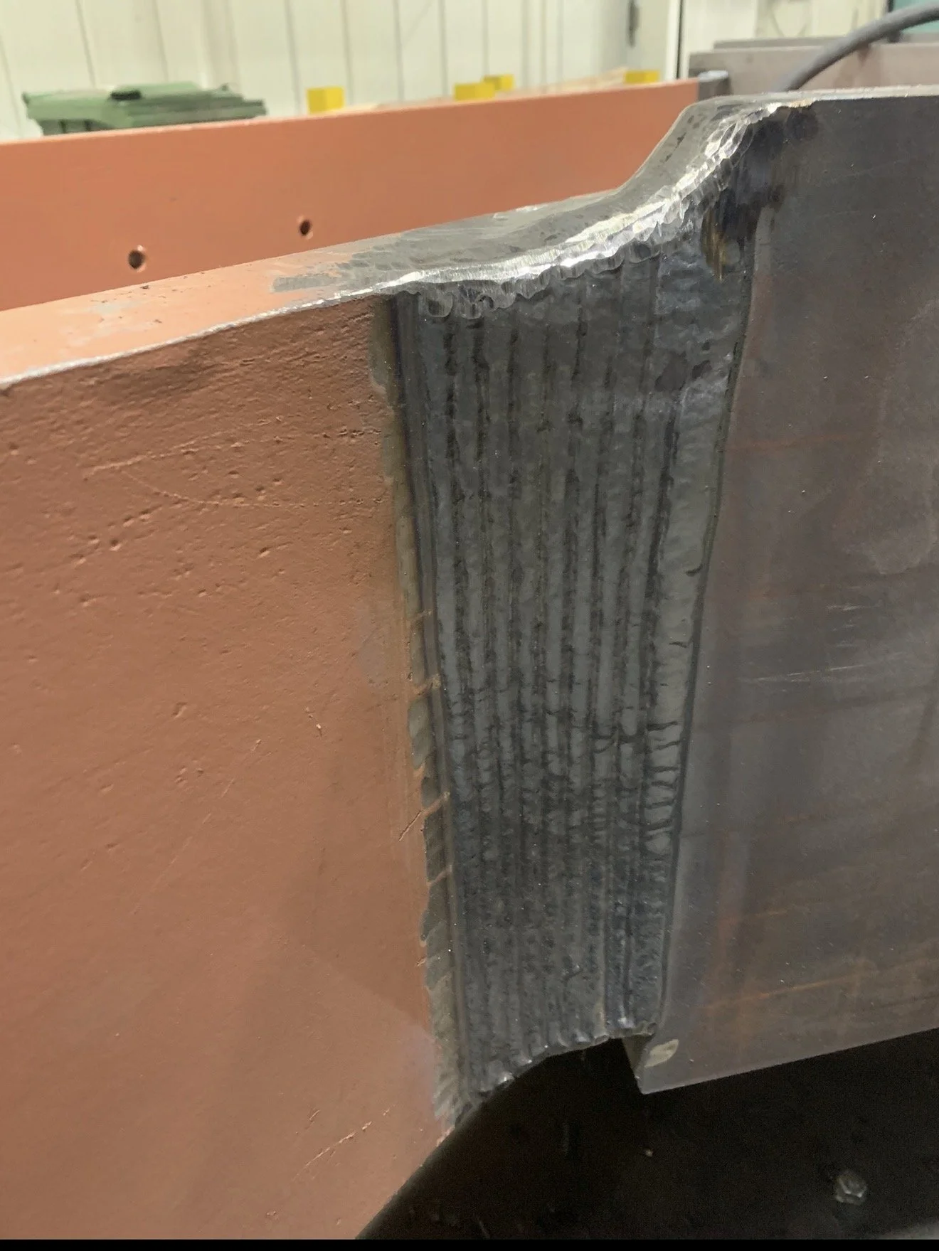 Close-up photograph of a metal corner joint. The image shows a metal piece welded to another with visible welding bead, with one piece painted orange and another unpainted gray metal. The setting appears to be a workshop or industrial environment.