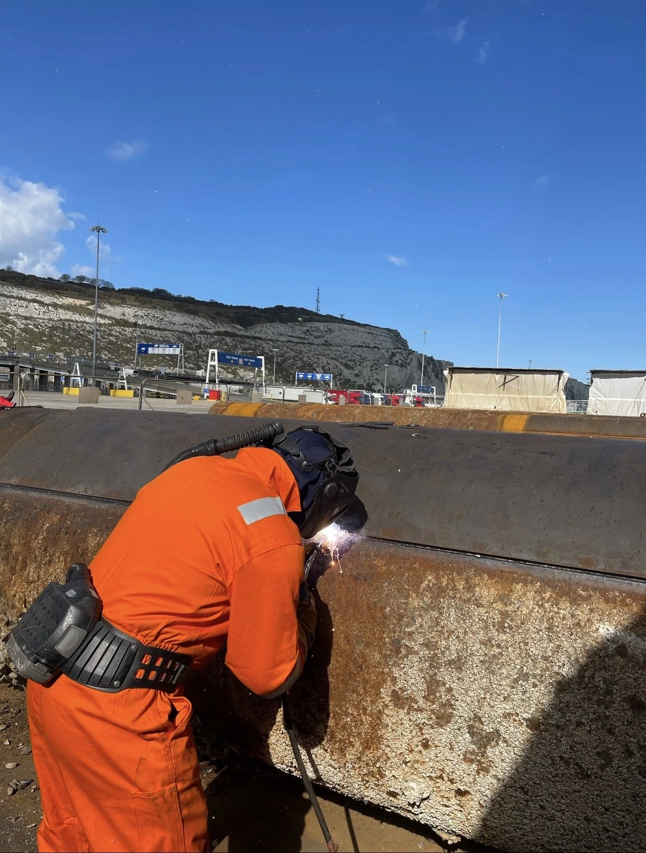 A worker in an orange safety suit is welding a large, rusty steel beam outdoors, with mountains and a clear blue sky in the background.