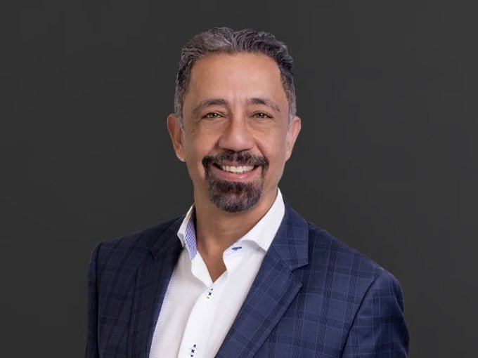 A smiling man with dark hair and a beard, wearing a navy blue checkered suit jacket and a white shirt, against a dark background.