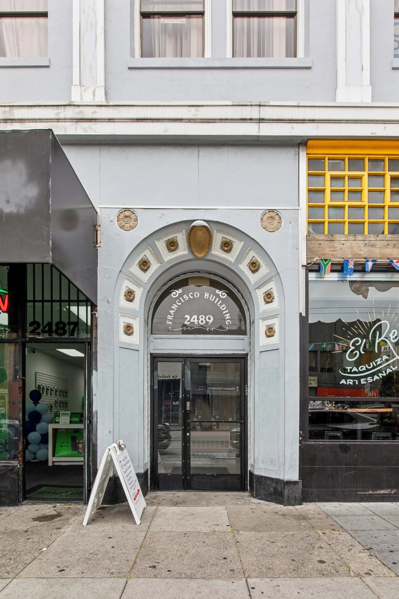 Facade of a building with an arched entrance labeled "Francisco Building 2489" and a glass door. To the left, there is an open storefront with balloons inside, and to the right, a store with flags and signage reading "EleP" and "artesanal."