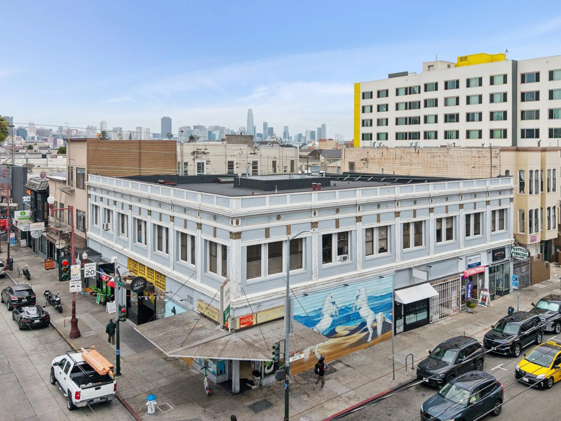 City street view showing multi-story buildings, storefronts, and parked cars, with a clear sky and the downtown skyline in the background.