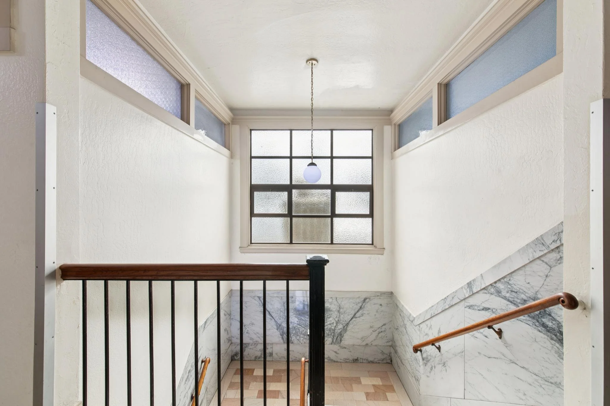 Interior view of a staircase landing with a large window at the end, a hanging ceiling light, marble flooring, and a wooden handrail on the staircase.
