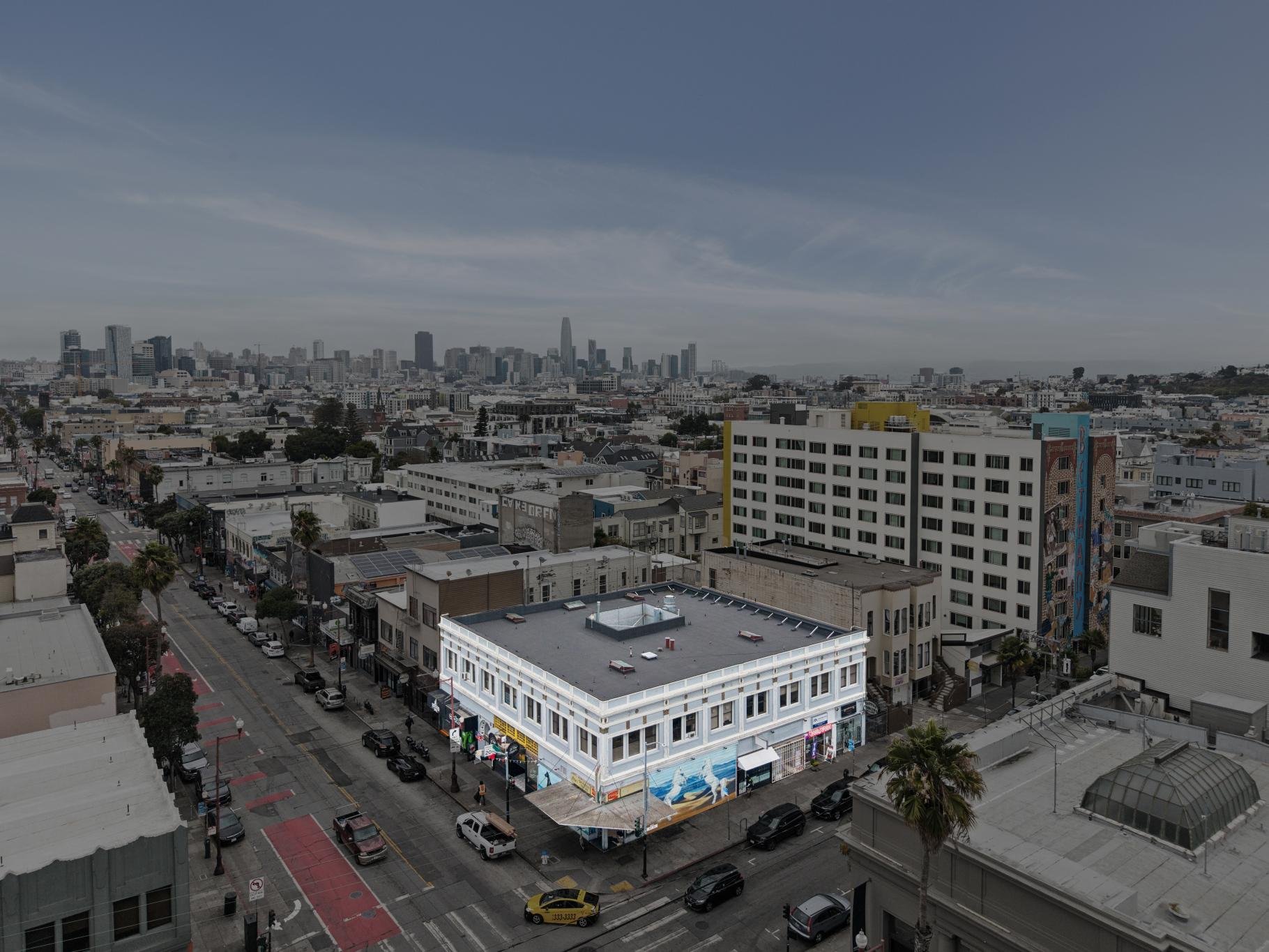 Cityscape of San Francisco showing buildings, streets, and a distant skyline including the Salesforce Tower under a partly cloudy sky.
