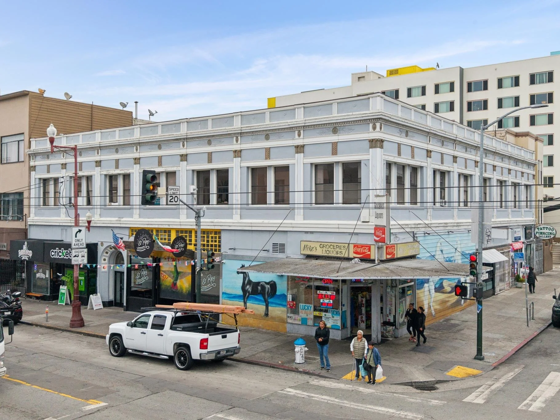 Street corner in an urban area with a two-story building housing a liquor store named 'Mike's Groceries Liquors,' with a mural of a cow on the exterior, near a traffic light and people walking on the sidewalk. A white pickup truck is parked nearby.