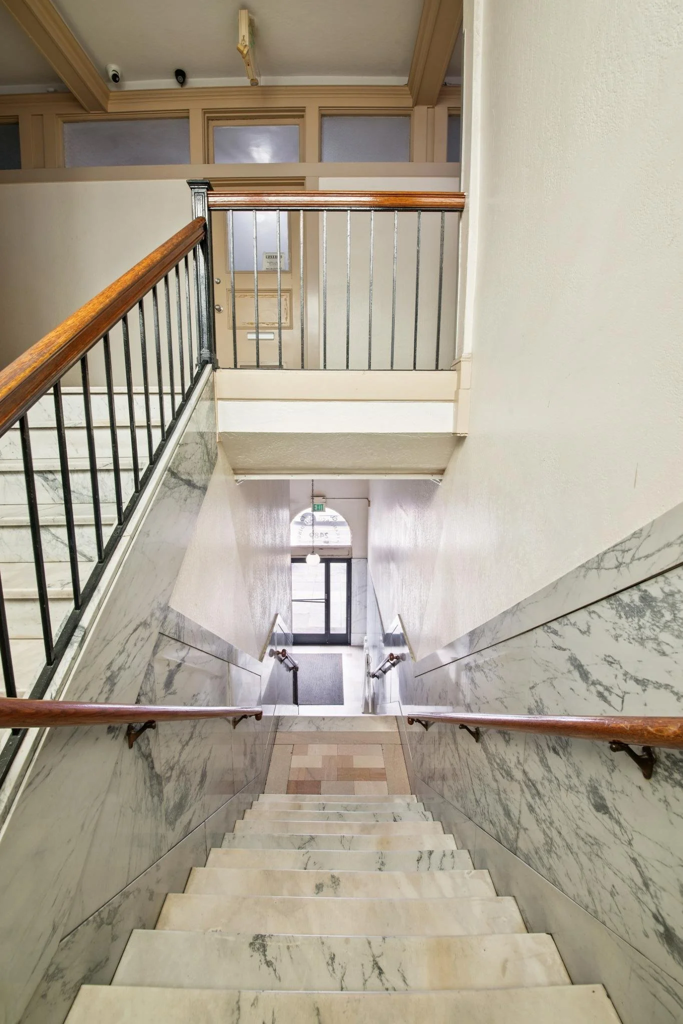 View of stairwell in a building with marble walls, beige steps, and wooden handrails, leading down to a door with an exit sign.