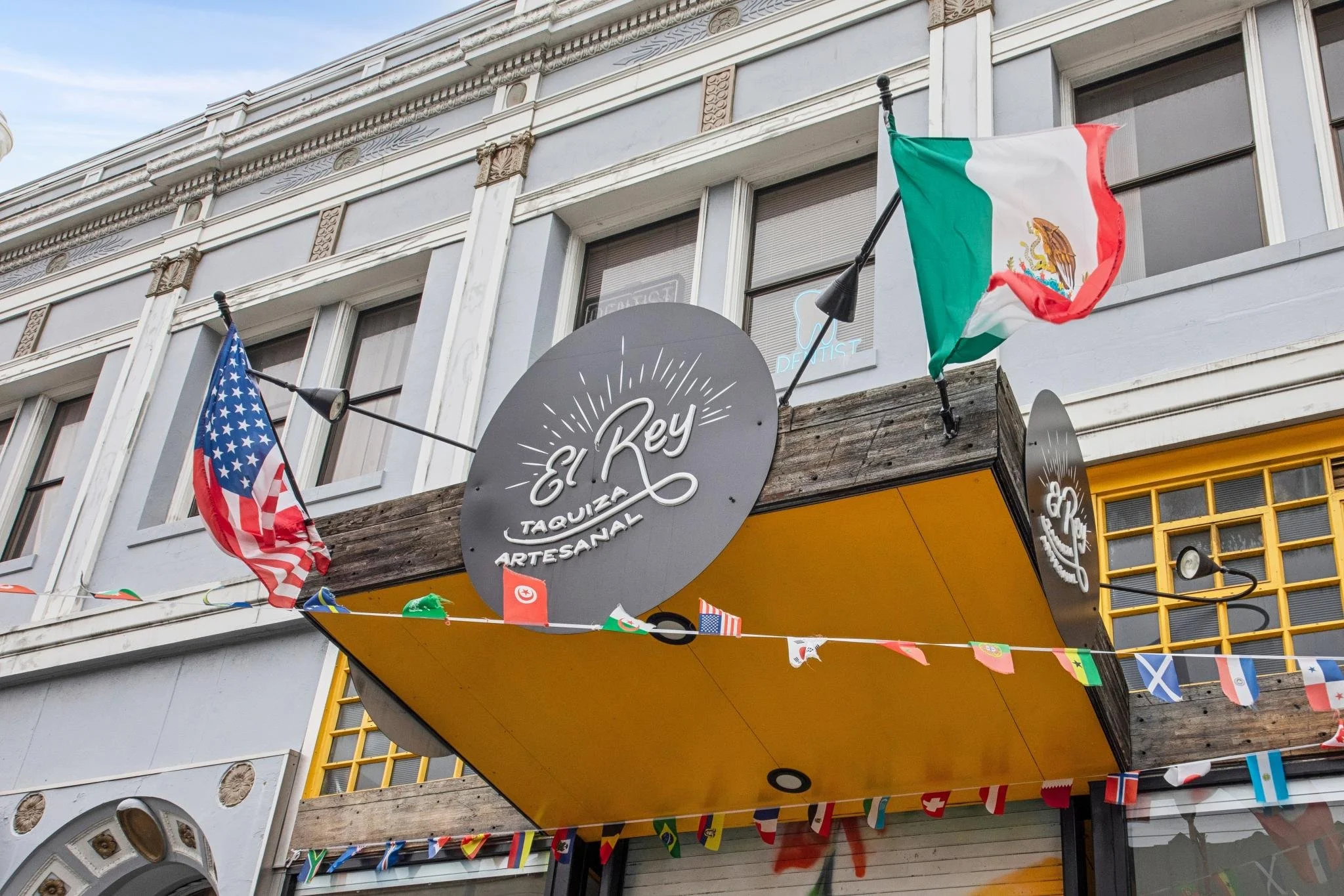 Building exterior with flags of the United States and Mexico, colorful decorative banners, and a sign reading 'El Rey Taqueria Artesanal'.