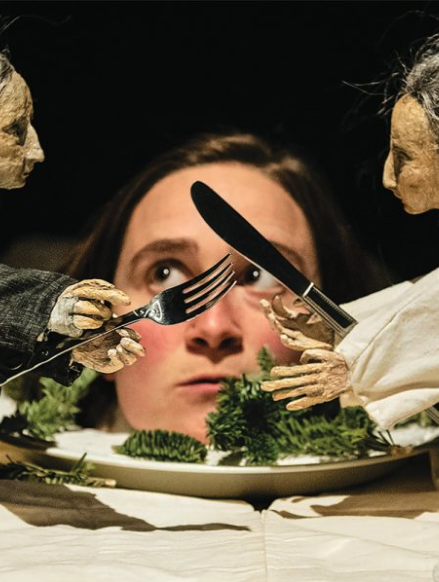 A woman with dark hair and a serious expression looking upward at two skeleton-like figures holding utensils and a knife and fork over a plate of greens.