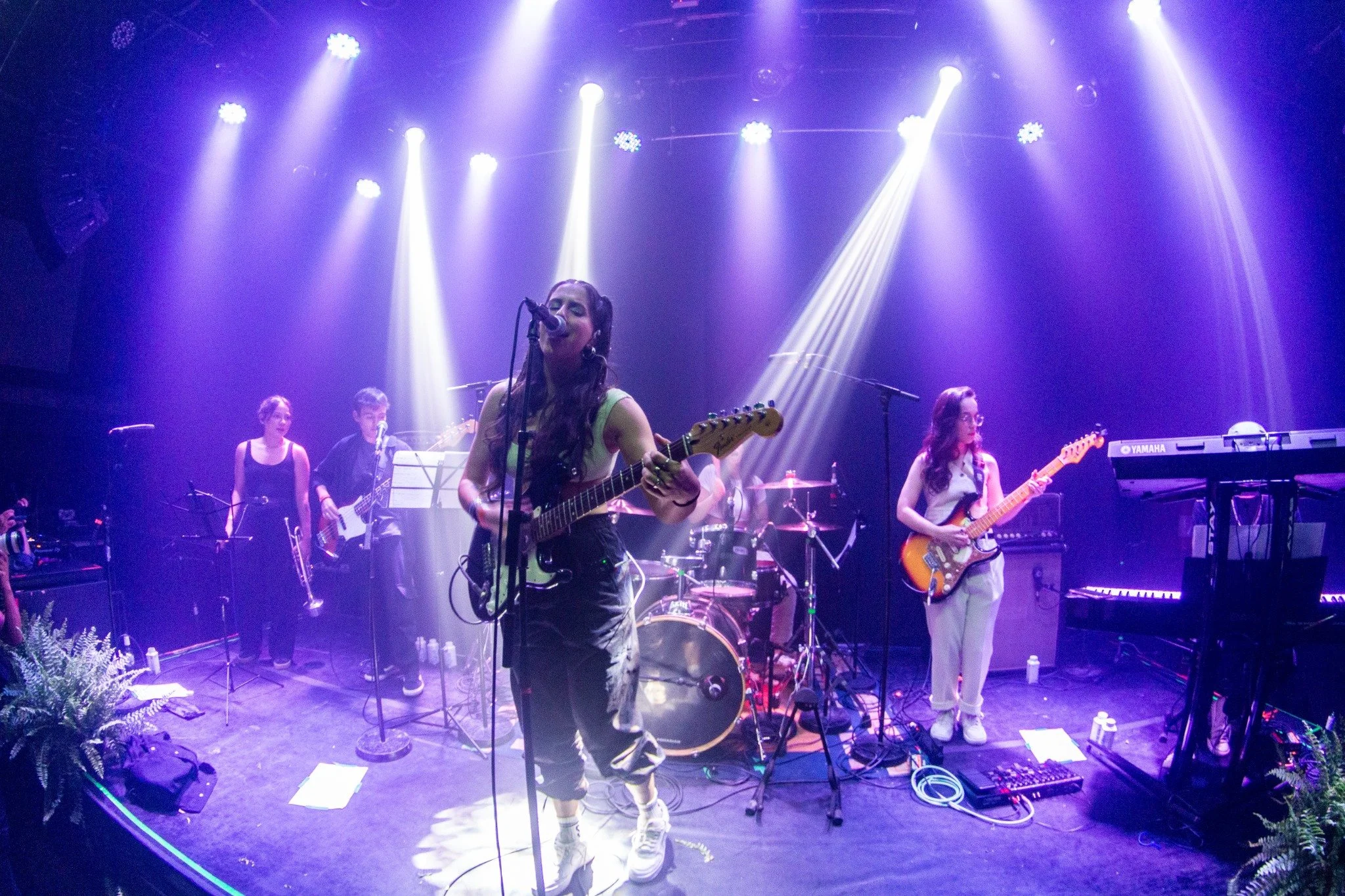 Pris and her band at the stage in one of her concerts, performing under purple stage lights. Pris is playing an electric guitar and singing into a microphone. Other band members are playing trumpet, bass, drums and keys