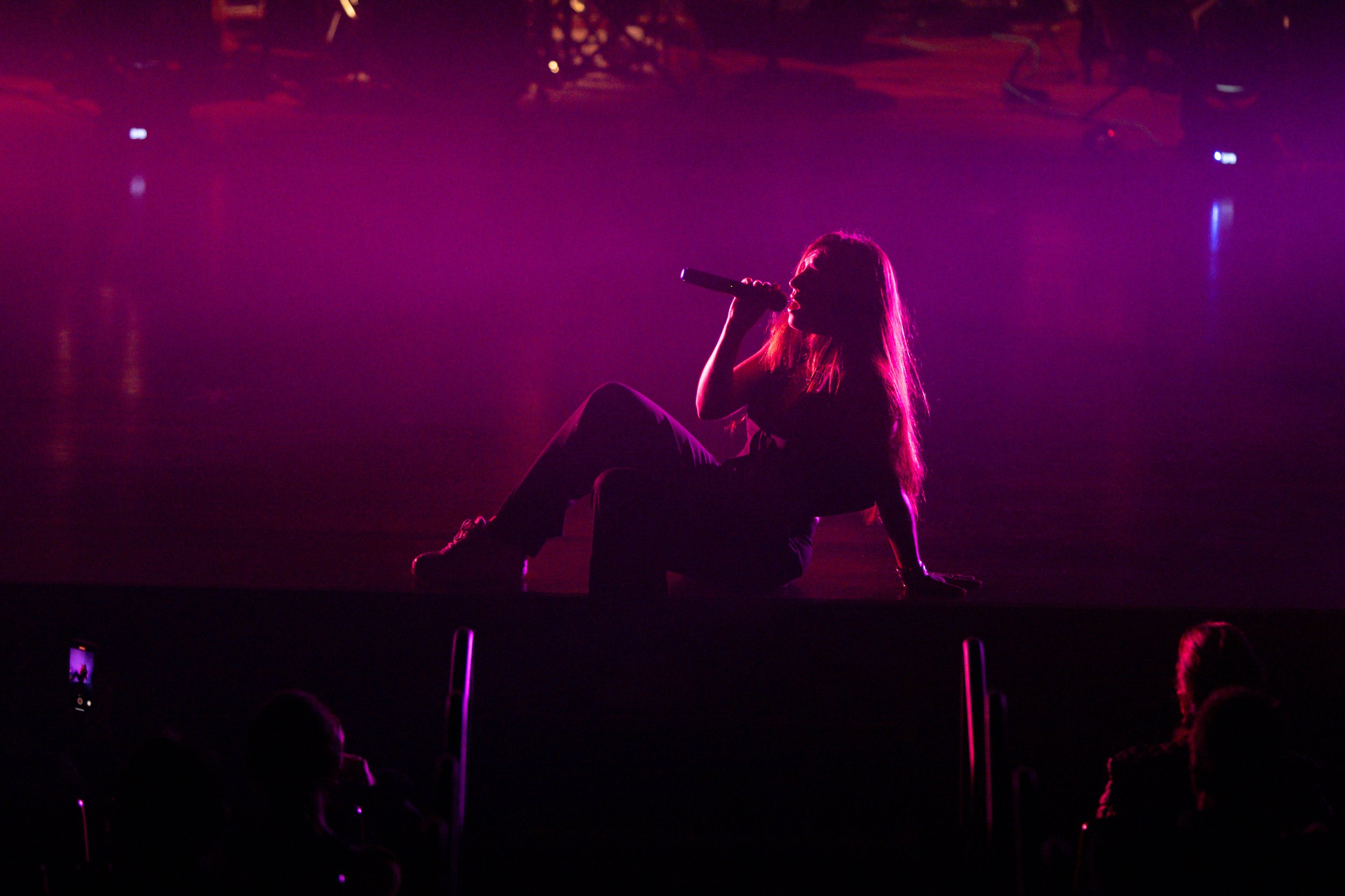 Pris sitting on the floor holding a microphone and singing in a dimly lit, purple-hued stage setting.