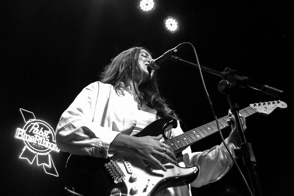Pris playing an electric guitar and singing into a microphone on stage, with a neon sign that reads "Pabst Blue Ribbon" in the background.