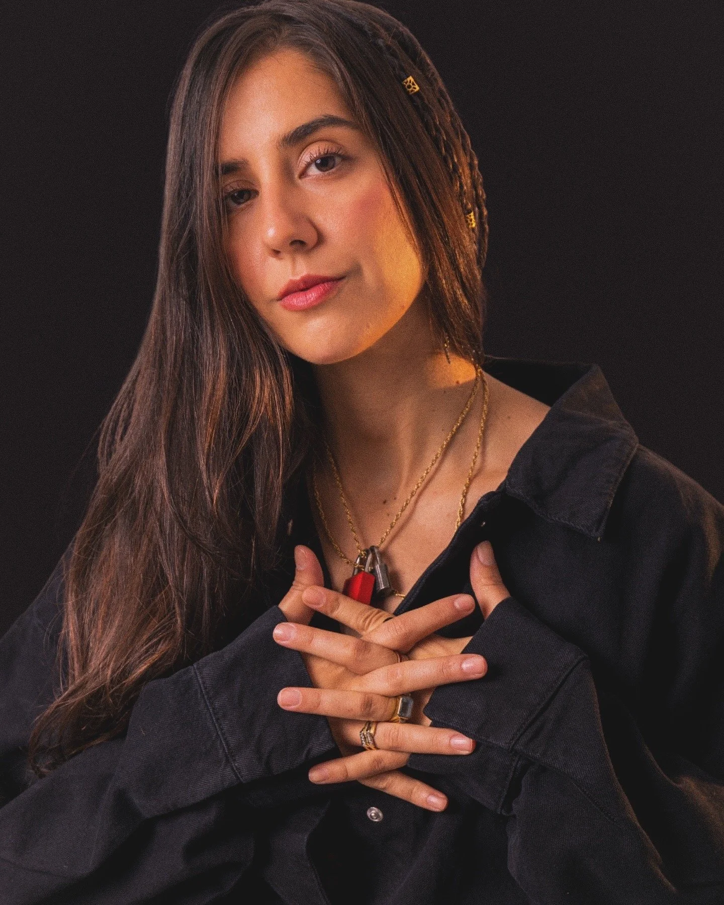Pris, a young woman with long brown hair and light skin in a black jacket, wearing gold jewelry and multiple rings, poses against a dark background with her hands crossed over her chest