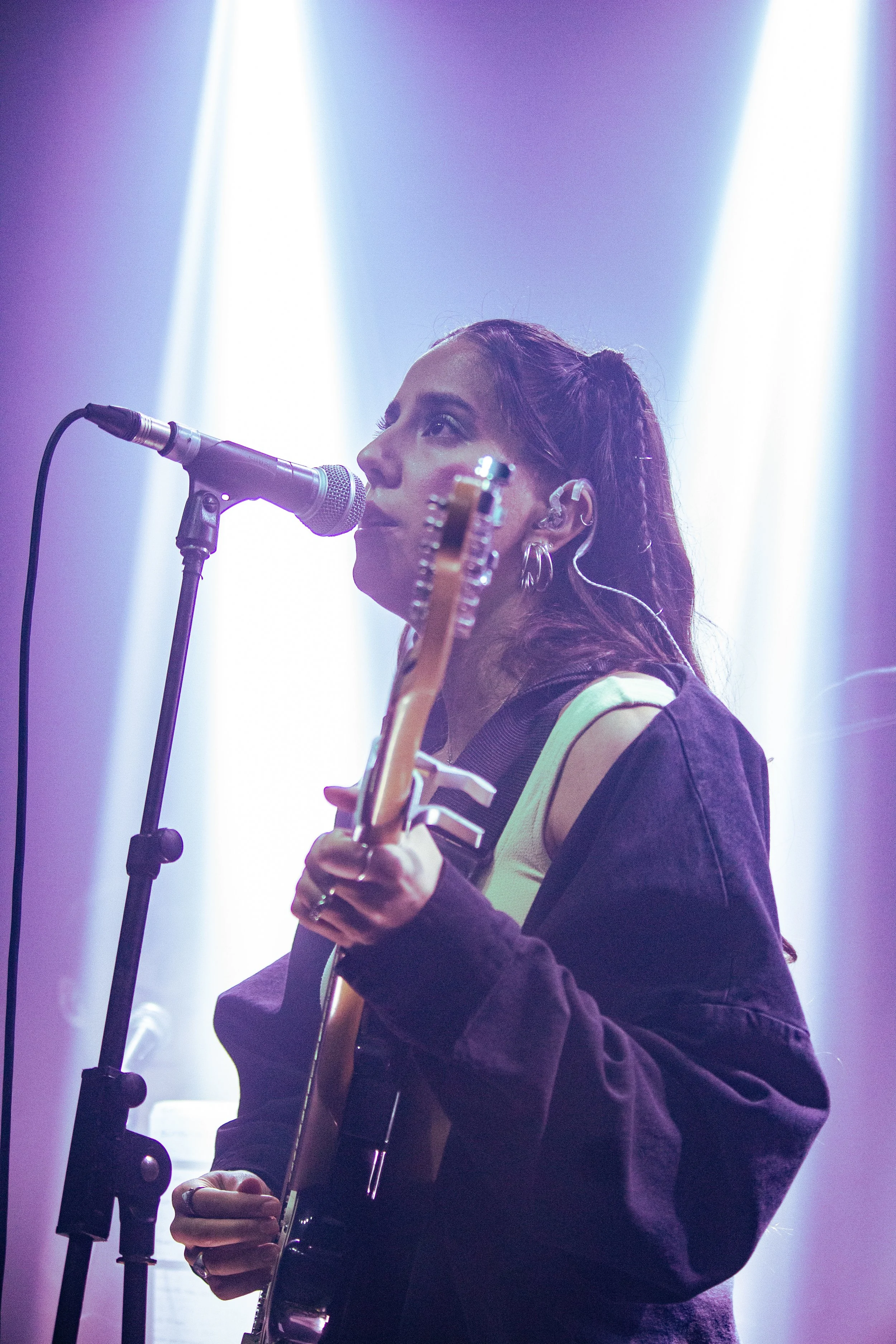 Pris, a woman with long, wavy hair playing an acoustic guitar and singing into a microphone on stage, illuminated by purple and white stage lights.