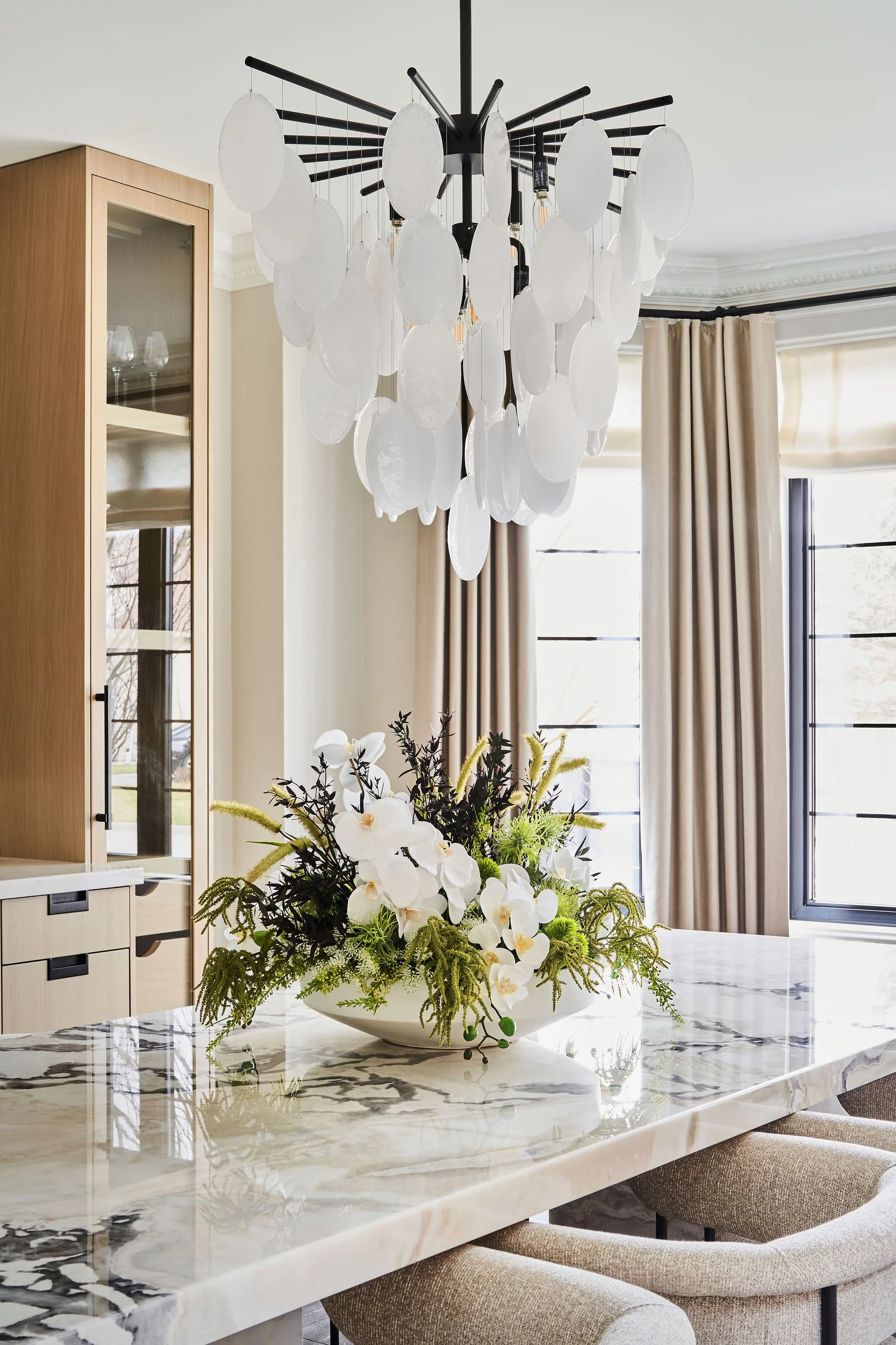 Modern dining room with a white-marble table, floral centerpiece, and chandelier with white, circular elements.