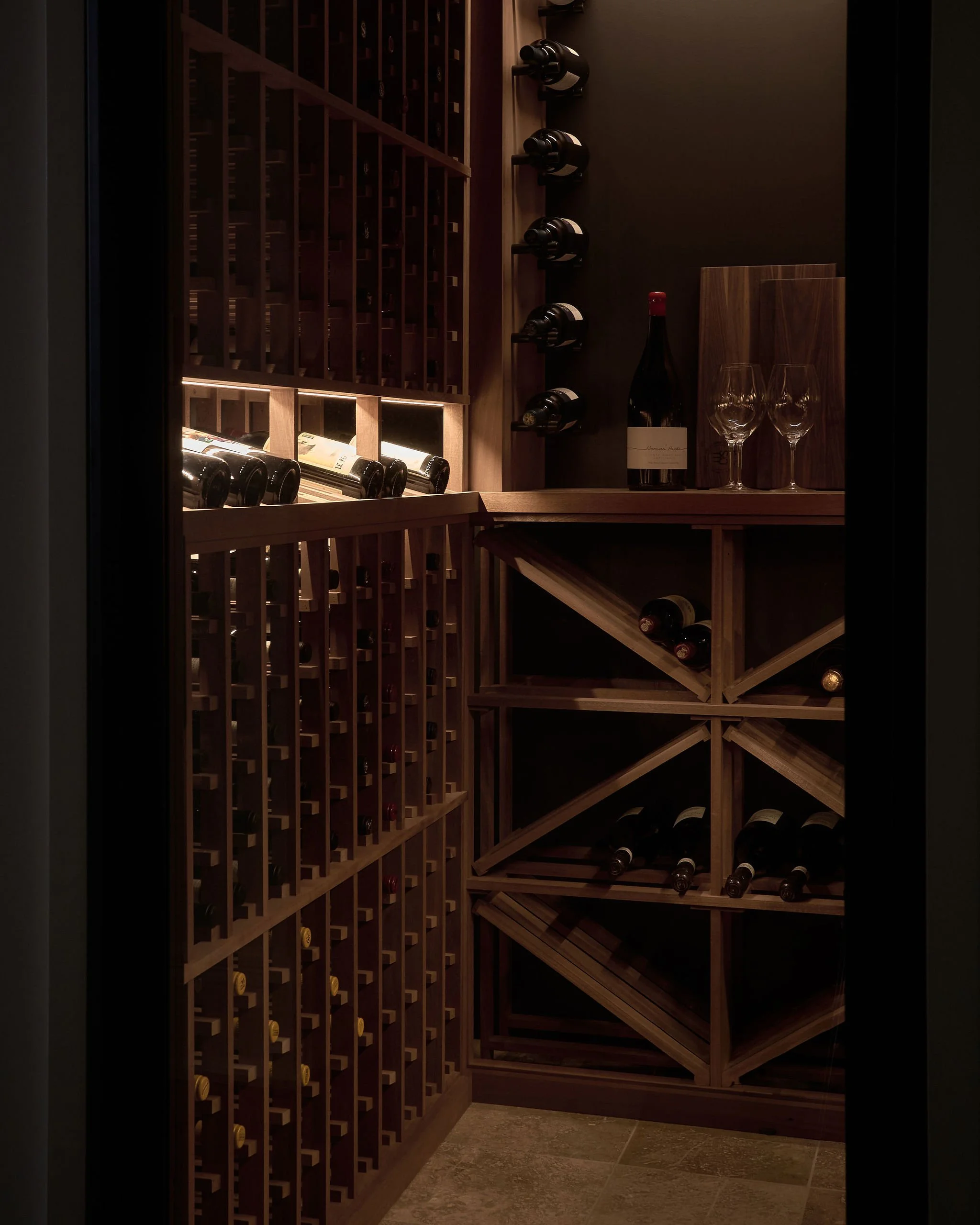 A wine cellar with wooden shelves filled with bottles, a bottle of wine with two empty wine glasses, and wine openers on the shelves.