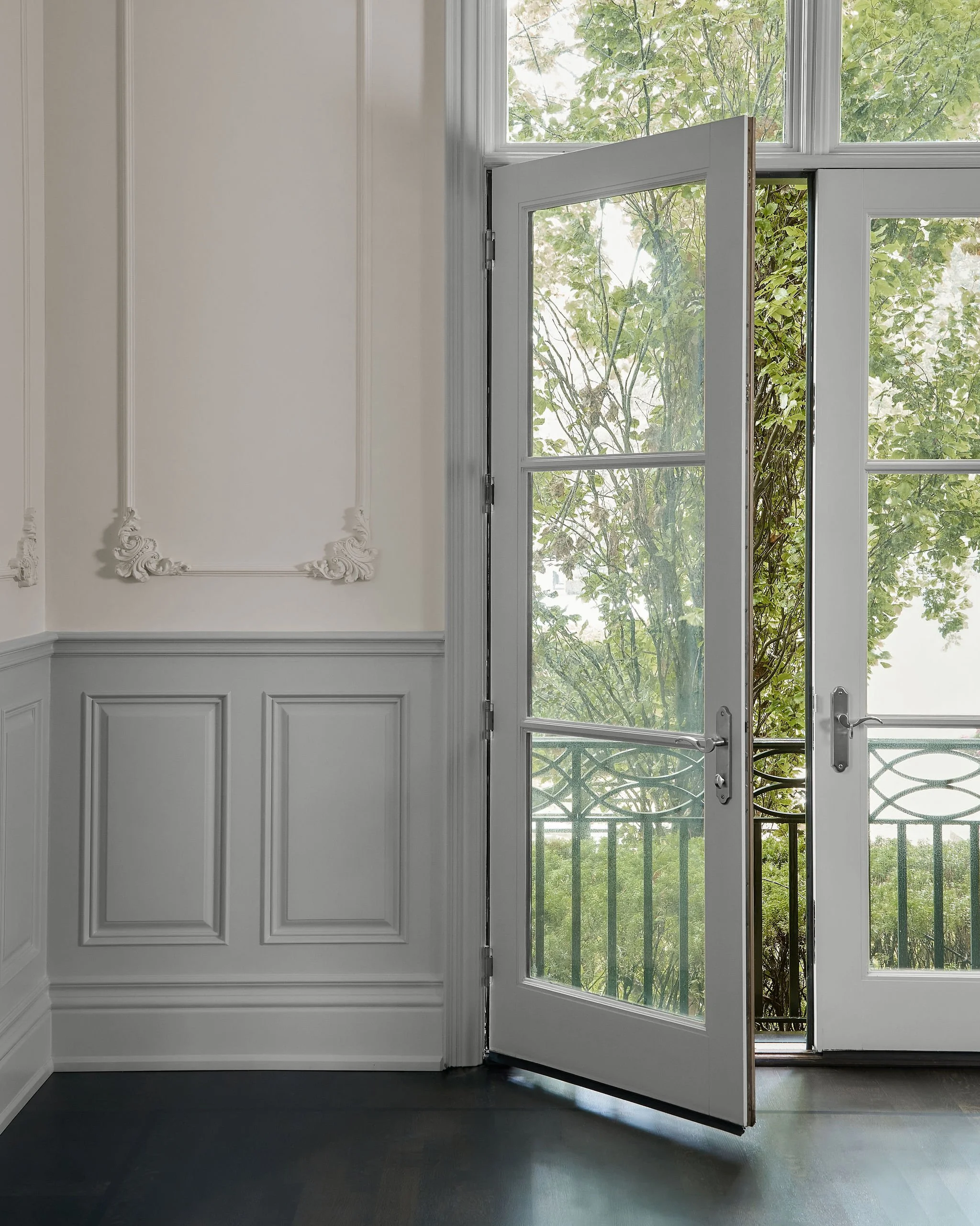 A white interior wall with decorative molding and a large glass door with white trim, partially open, showing a balcony with a black railing and green trees outside.