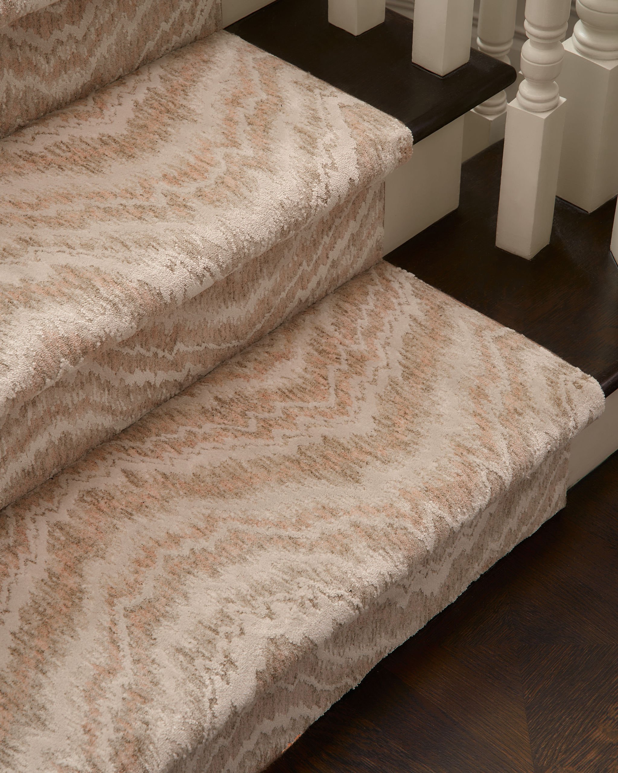 Close-up of a beige patterned carpeted staircase next to a dark wooden floor and white decorative balusters.