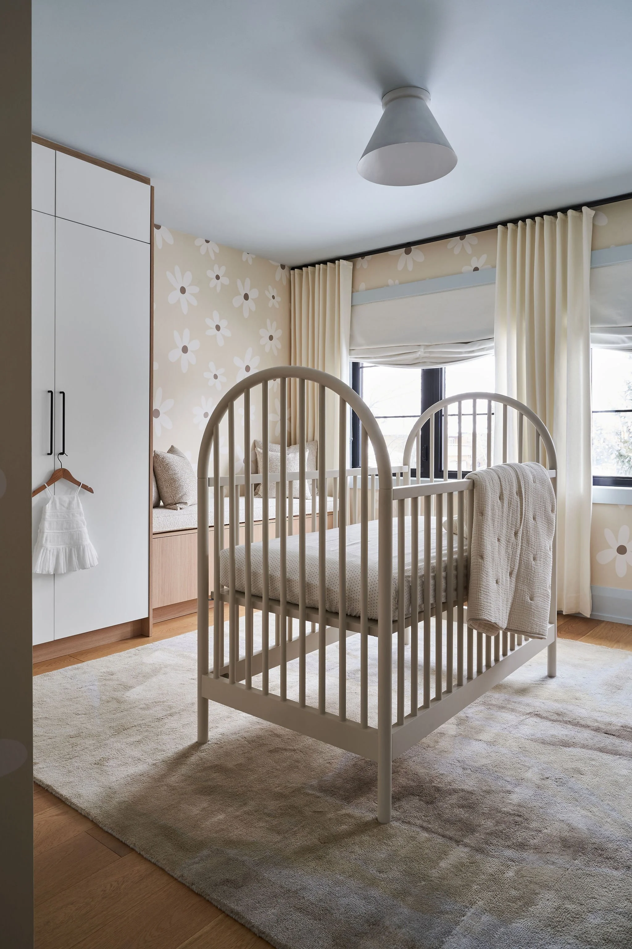 A cozy nursery with white crib, beige floral wallpaper, large window with cream curtains, white built-in wardrobe, and beige area rug.