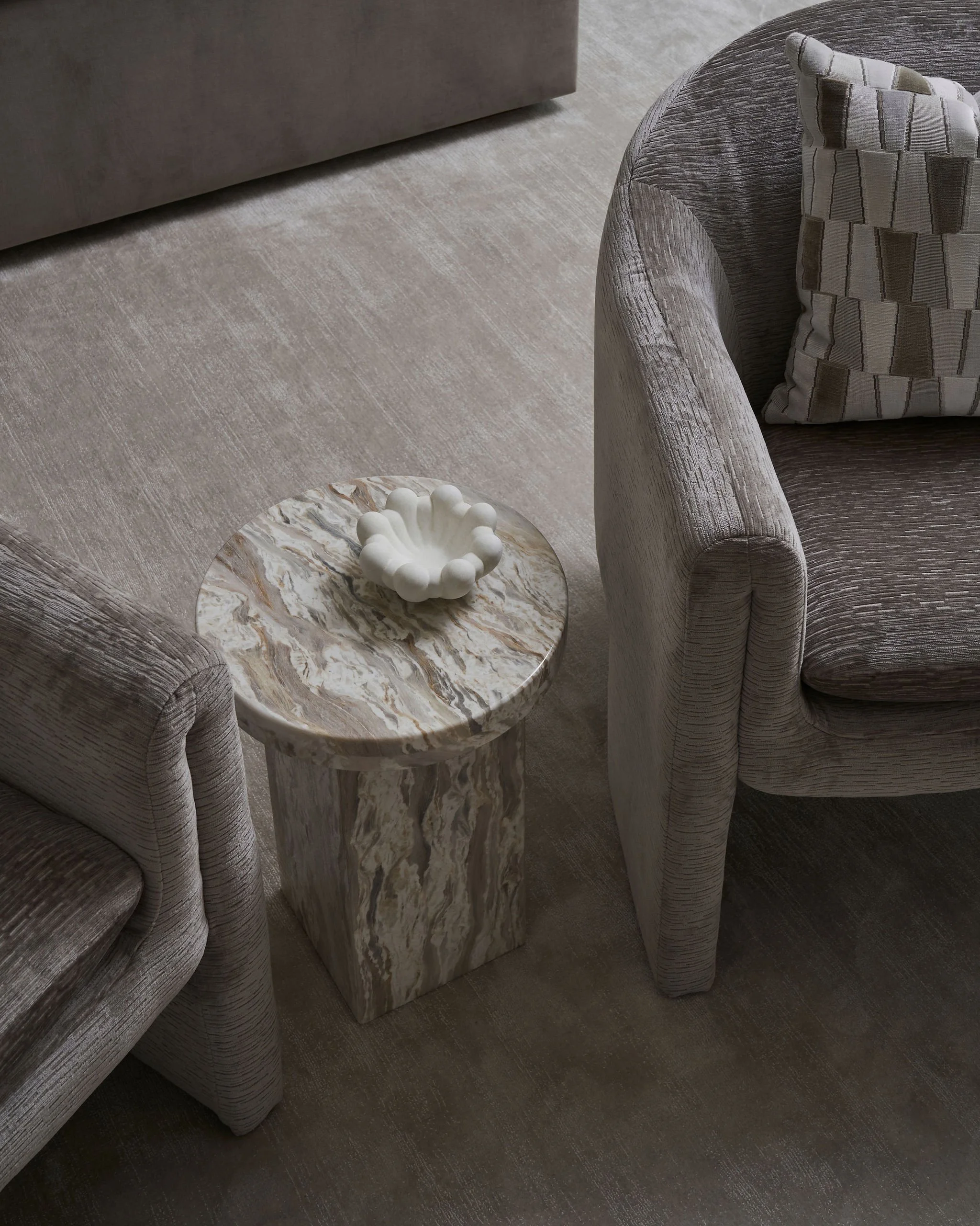 Two upholstered armchairs with rounded arms and a patterned throw pillow on one of them, positioned around a marble-top side table with a decorative white ceramic bowl on top, situated on a beige carpeted floor.