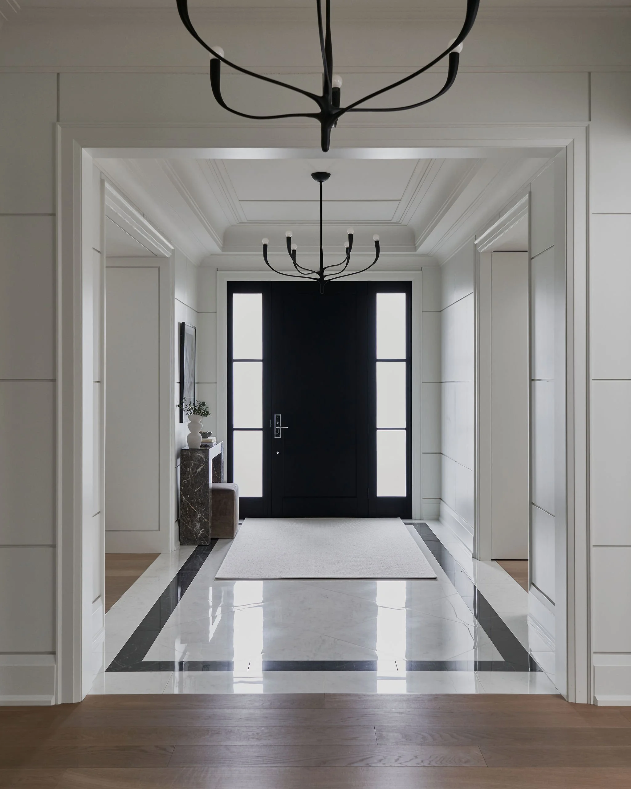 Elegant entryway with black front door, white walls with panel molding, black chandelier, marble flooring with black and white border, small side table with plant, and wooden floor in an adjoining room.