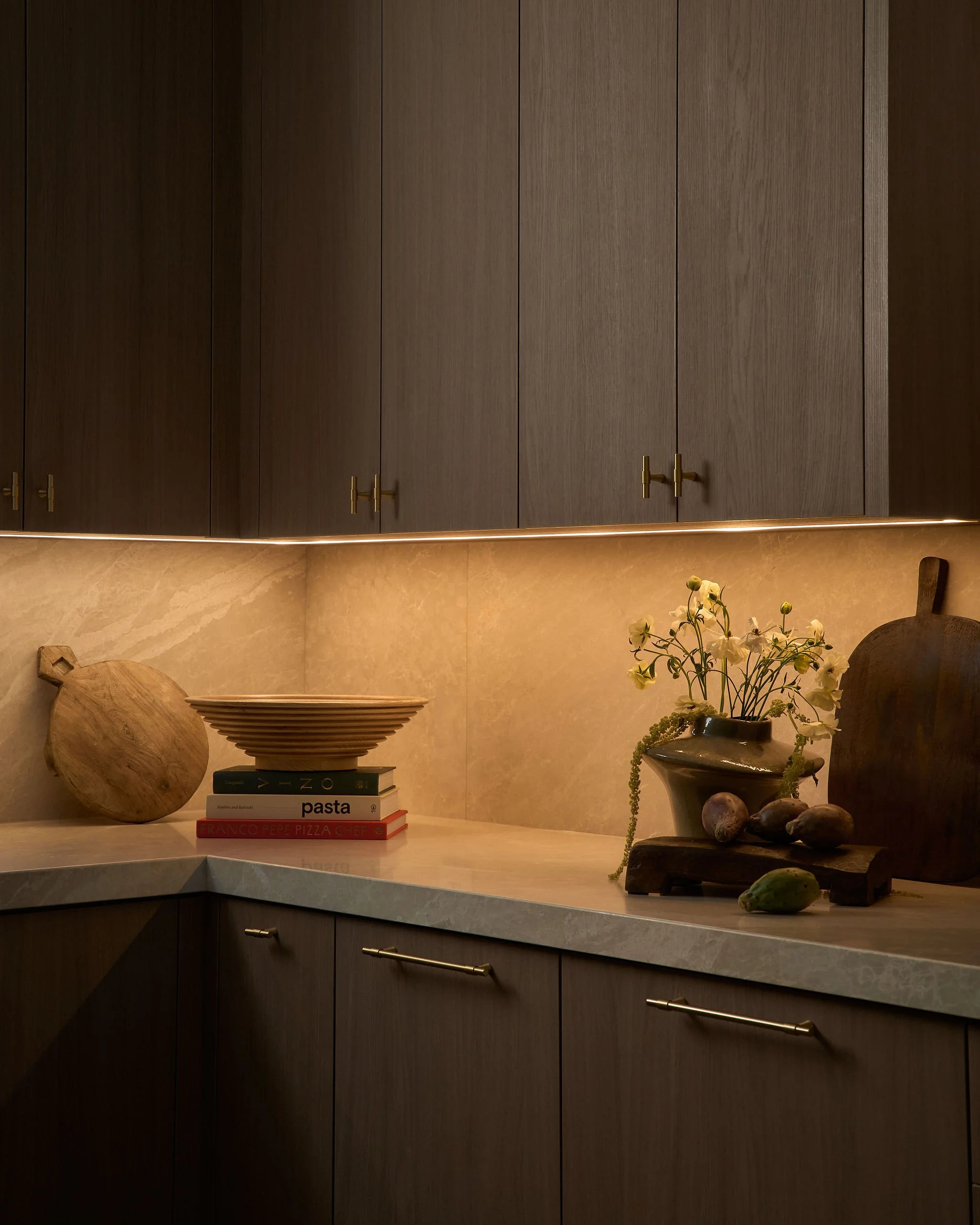 A kitchen countertop with a wooden cutting board, a stacked set of cookbooks, a decorative bowl, and a vase with white flowers and green foliage, set against a wall with brown wooden cabinetry.