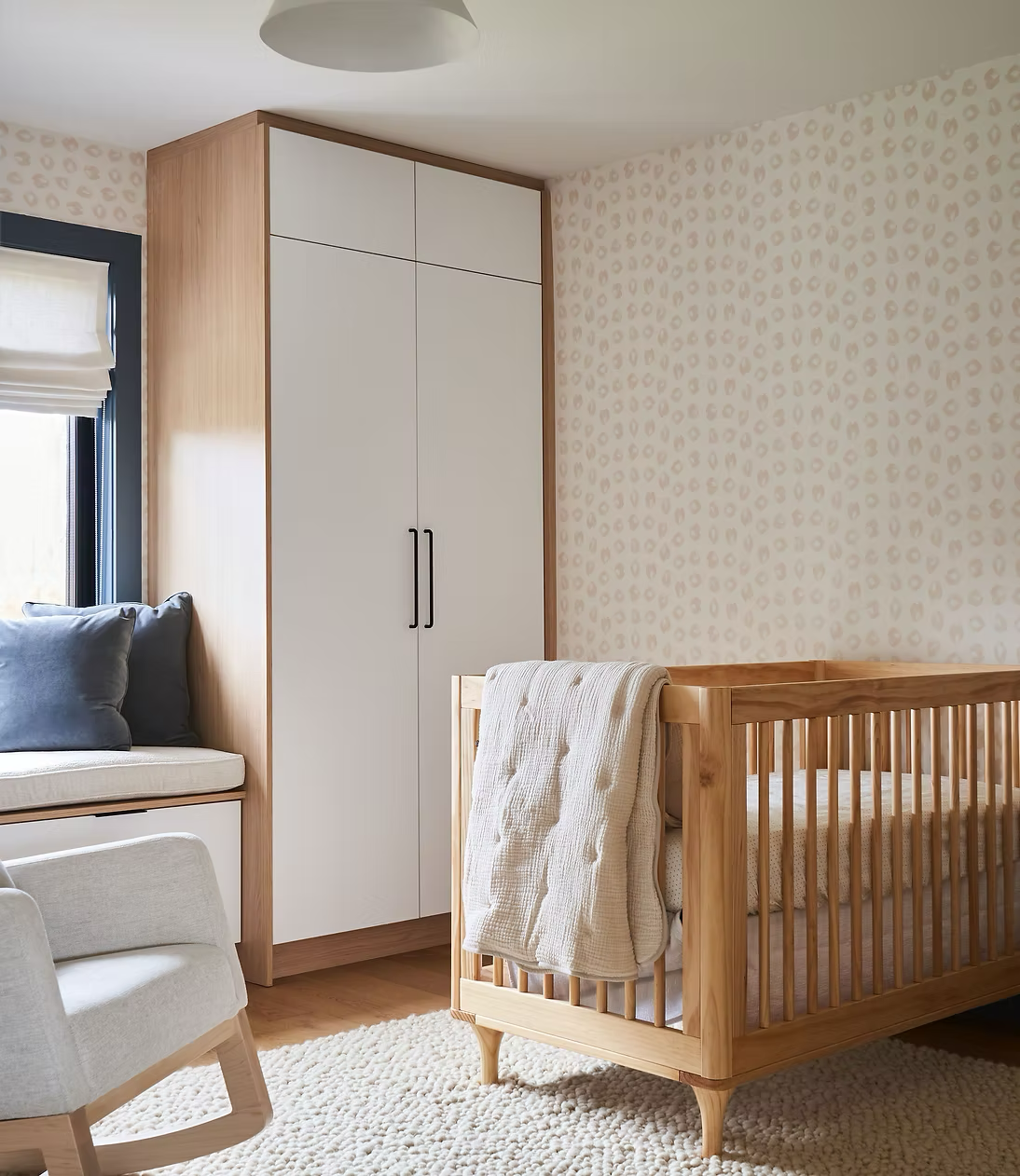 A nursery room with a wooden crib draped with a cream blanket, a white wardrobe with black handles, a window with beige blinds, a gray cushioned chair, and a beige textured wall with a subtle leopard print pattern.