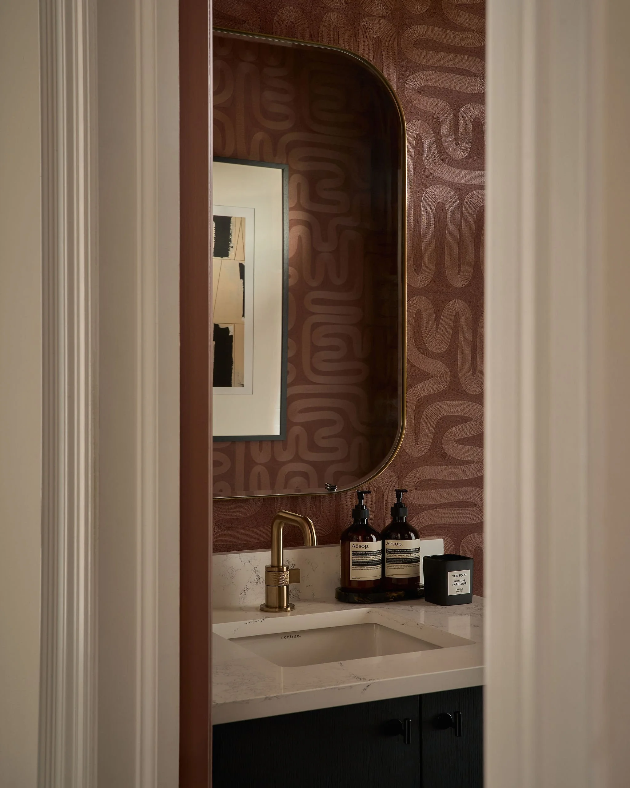 A bathroom vanity area with a white marble countertop, a white sink, a gold faucet, two brown bottles labeled 'Aesop', a round black candle labeled 'Tom Ford', and a large mirror above the sink. The wall behind features wavy, patterned wallpaper in s