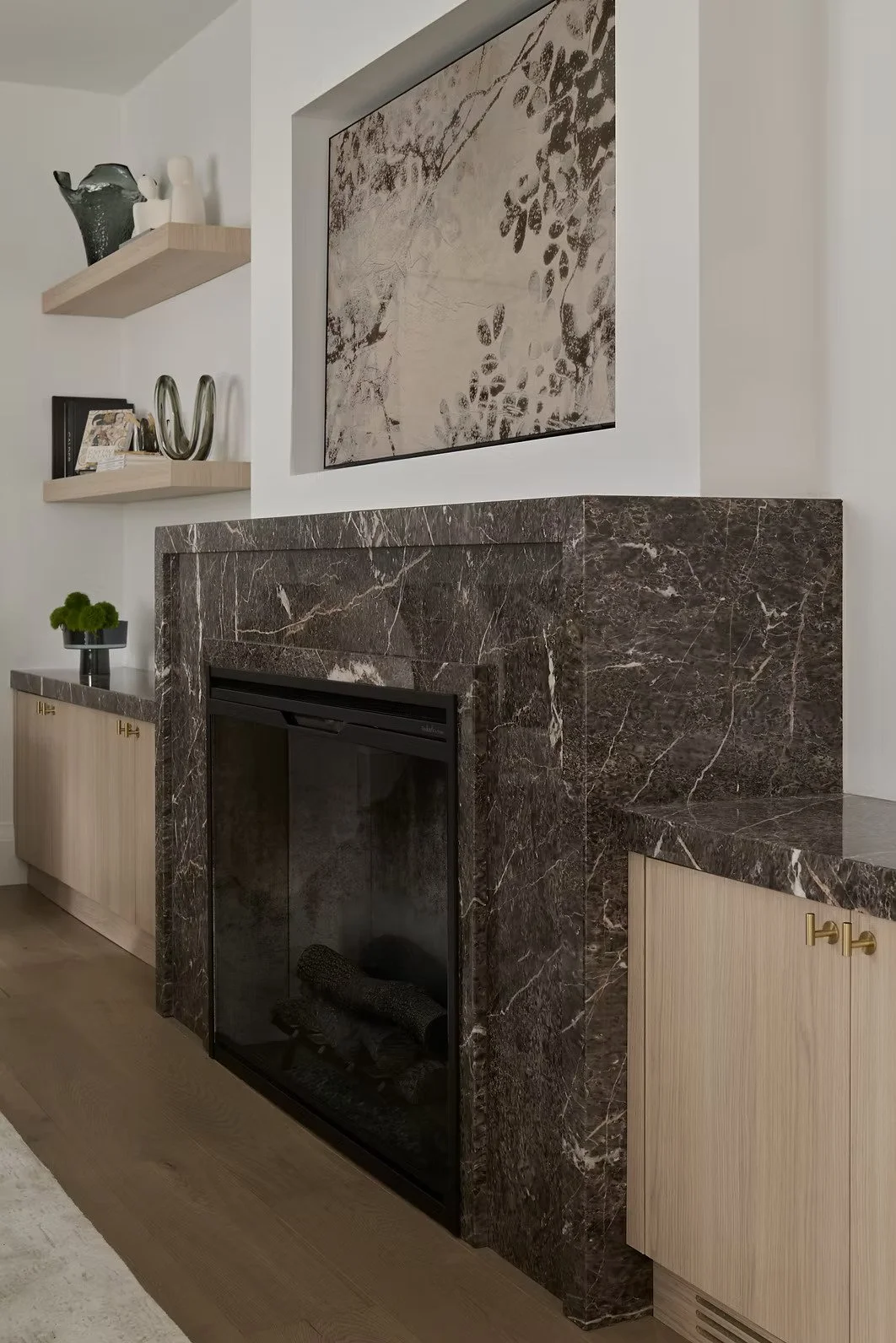 A modern living room fireplace with a dark brown marble surround, light wood cabinetry, a large abstract painting above, and decorative shelves with vases and books.