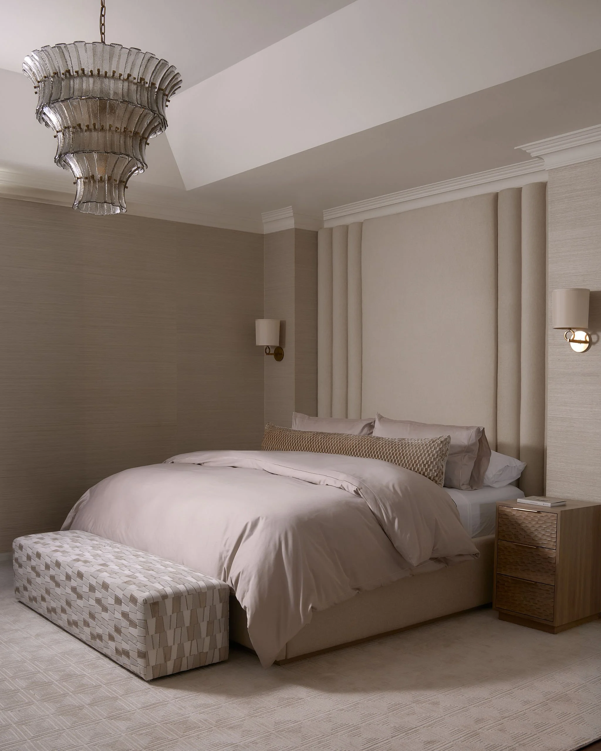A neatly made bed with beige linens and matching pillows in a bedroom with beige walls and patterned carpet. A nightstand with a wicker design is beside the bed, with wall-mounted lights on either side.