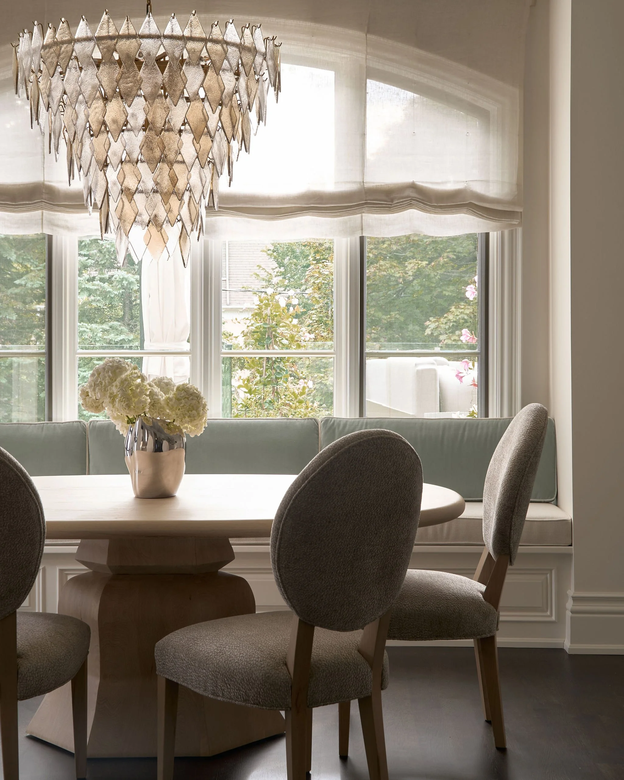 A dining room with a light wood round table, upholstered chairs, a vase with white hydrangeas, a window with white curtains, and a chandelier hanging from the ceiling.