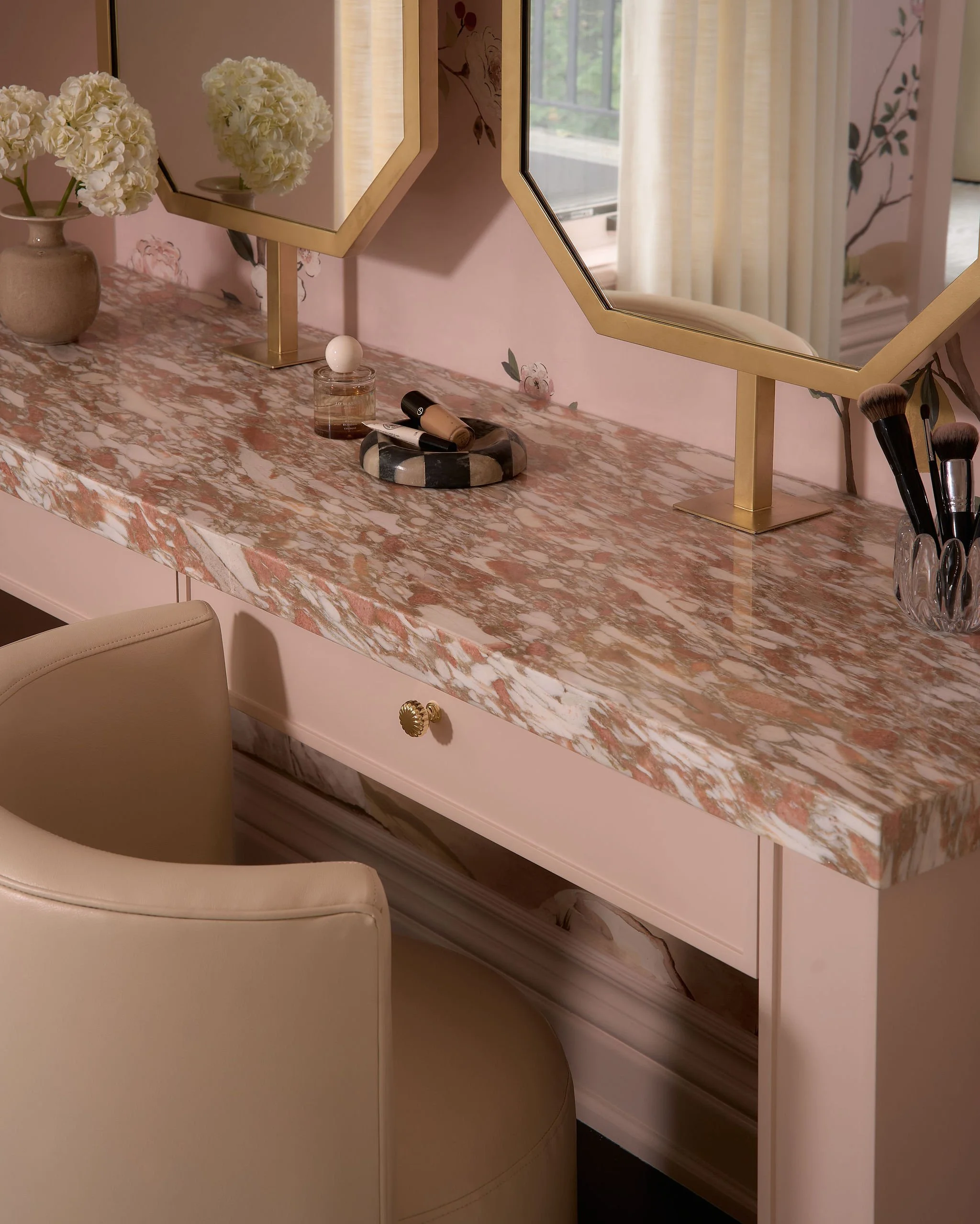 A marble vanity table with a pink, white, and orange pattern. Two hexagonal mirrors with gold borders are mounted on the wall above the vanity, reflecting nearby window curtains and window. The table holds a flower vase with white hydrangeas, a perfu