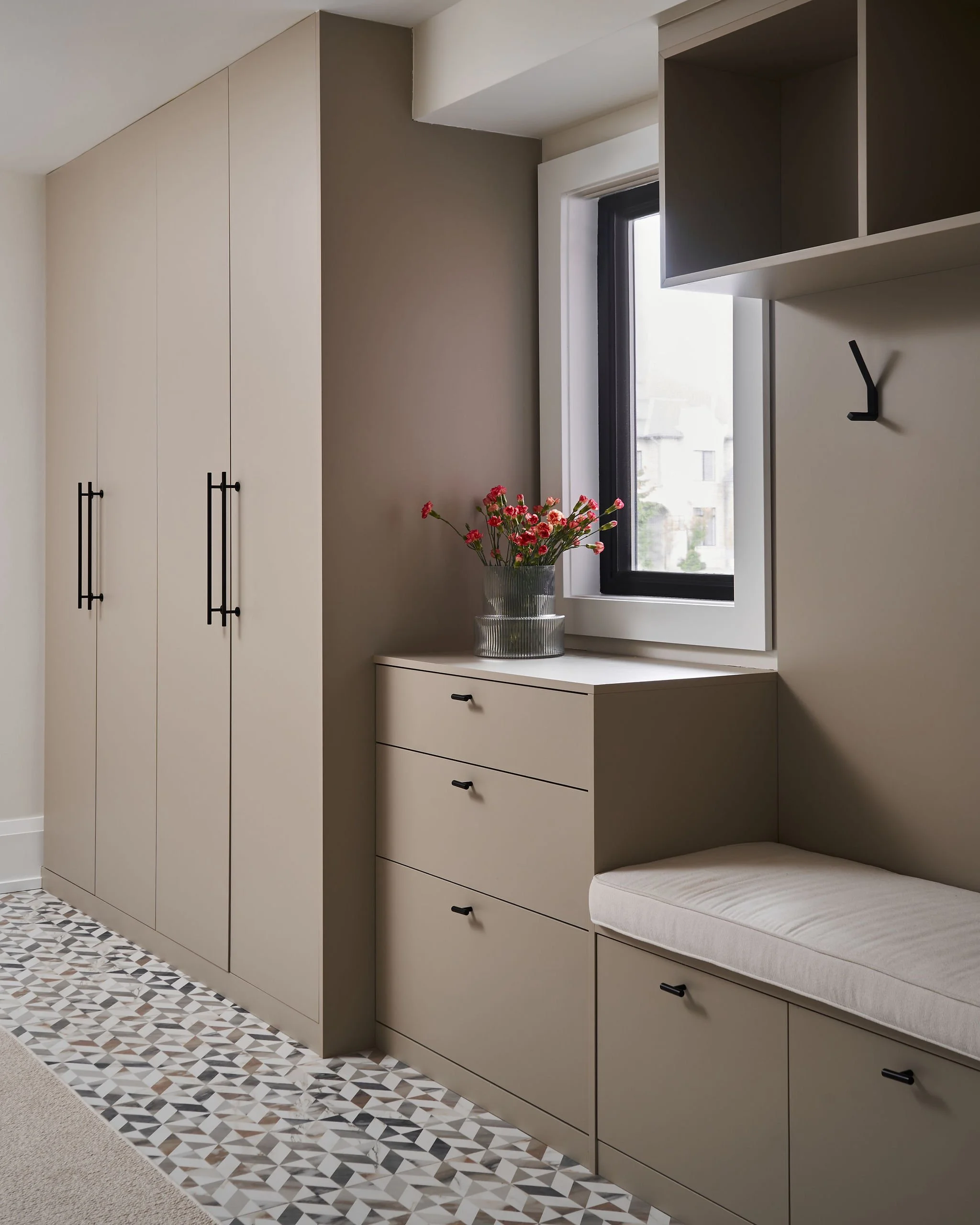 Modern kitchen corner with beige cabinets, a window, a bouquet of pink flowers in a glass vase, and a built-in seating area with a cushion, all set against a geometric patterned tiled floor.