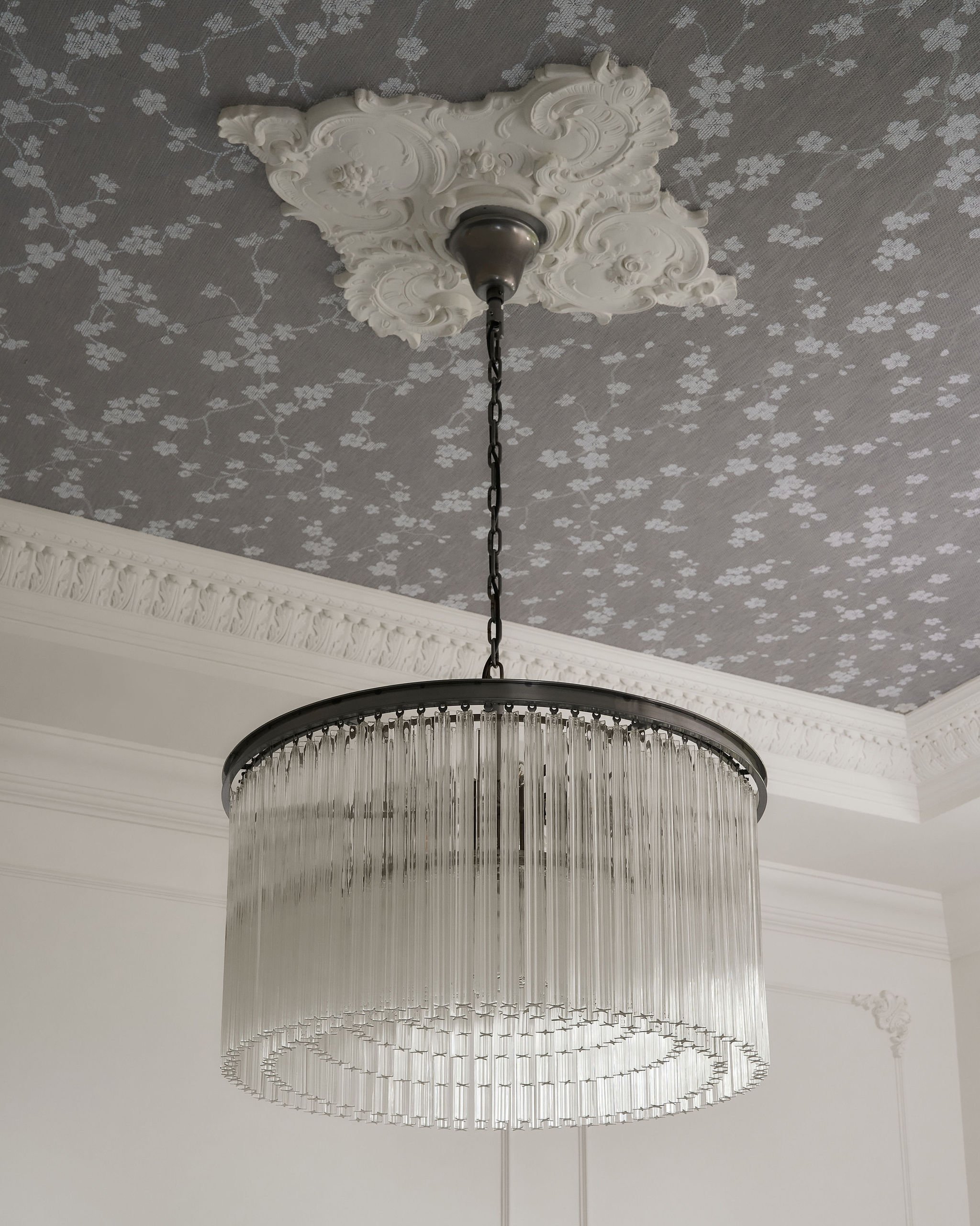 A large circular chandelier with hanging crystal-like rods, hanging from a ceiling with a decorative ceiling rose and floral patterned wallpaper.